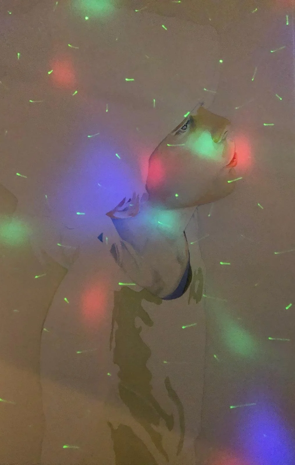 A cartoon-style drawing of a person with head tilted back and eyes closed, illuminated by colorful laser lights in red, green, and blue. The drawing appears to be on a wall, with reflections of the lights scattered around.