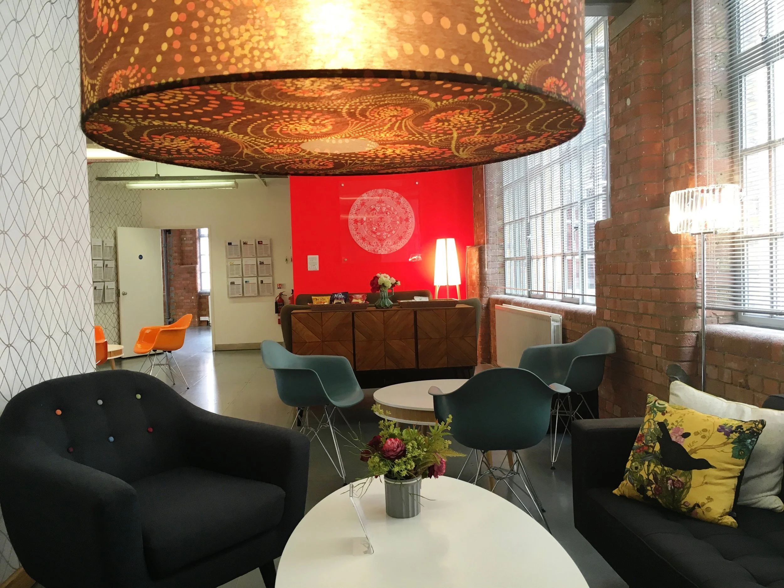 Interior of a modern office or lounge with brick walls, large windows, and various seating options including black, blue, and gray chairs. There are two white round tables, one with a flower arrangement. A large decorative lamp hangs from the ceiling