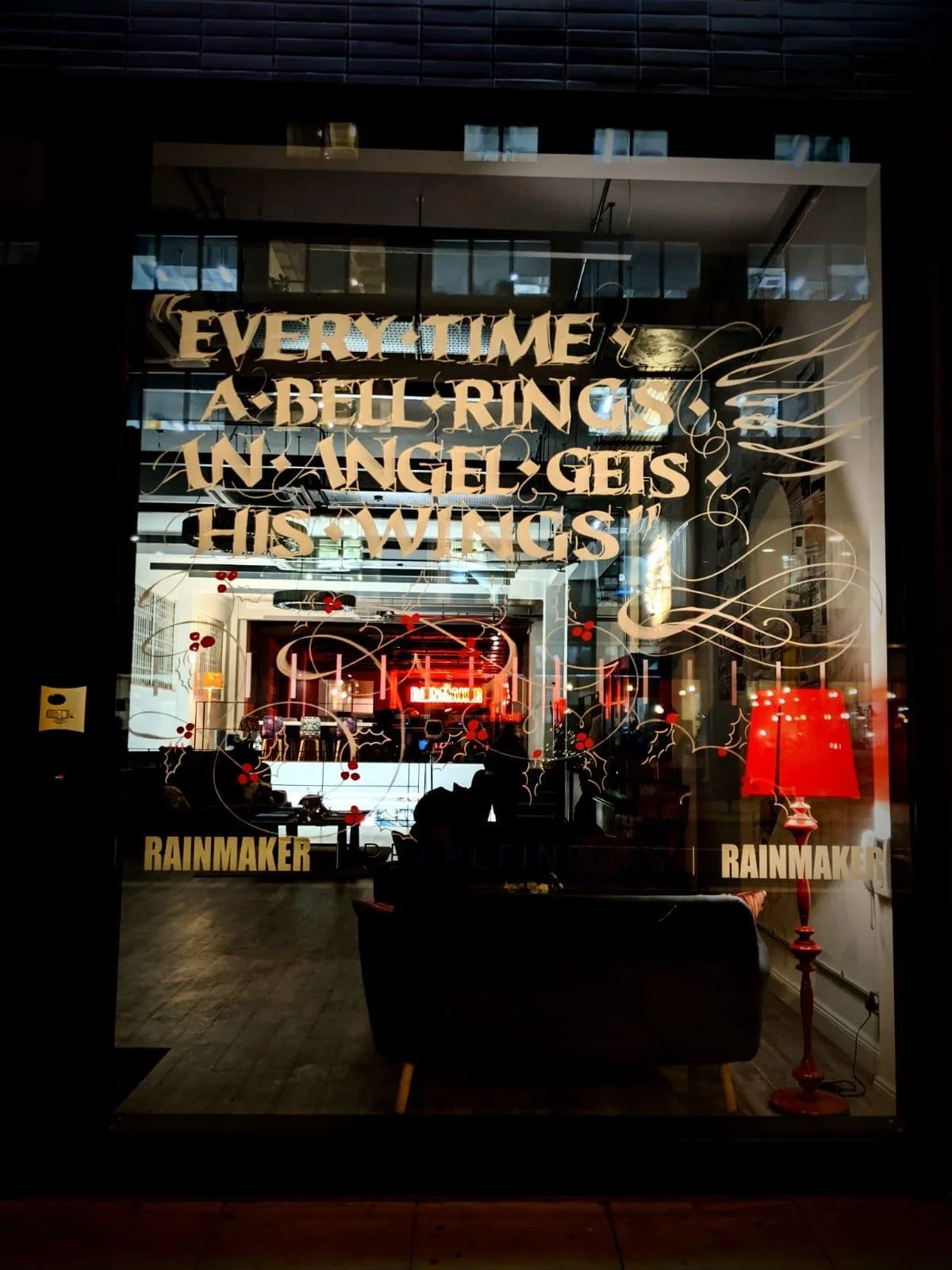 A glass window with gold lettering that reads "Every time a bell rings, an angel gets his wings." Inside, there is a red lamp, chairs, and a reflection of a lit neon sign that says "Rainmaker."