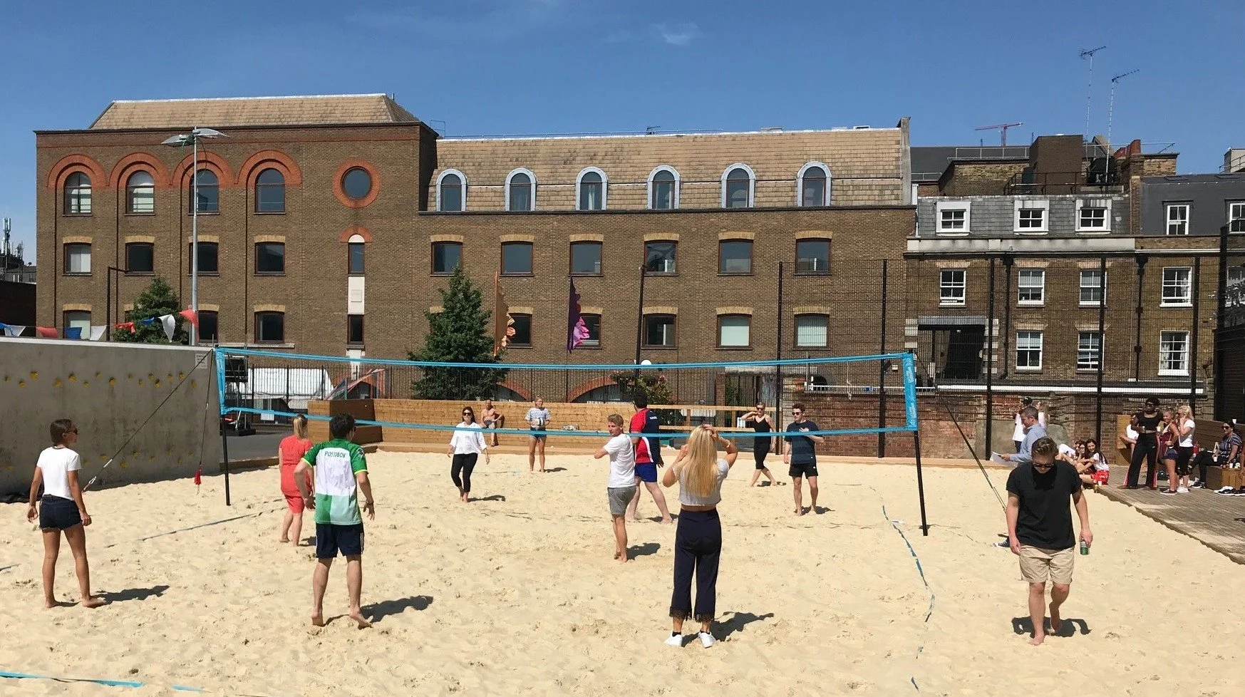 People playing beach volleyball on a court with sand, surrounded by buildings under a clear blue sky.