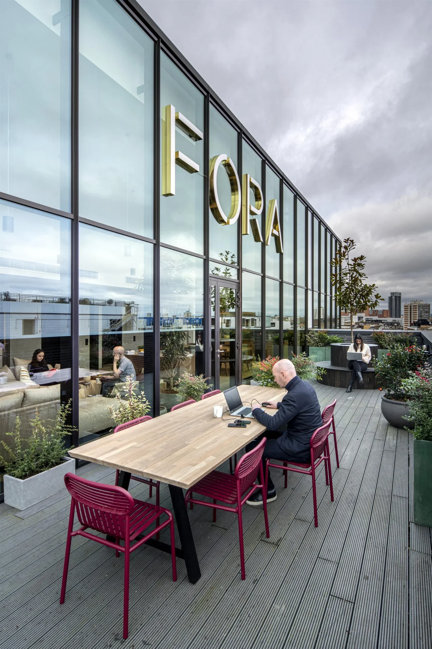 Rooftop outdoor workspace with a large wooden table and pink chairs, several people working on laptops, glass building with a sign that says 'FORA', and cityscape with tall buildings under cloudy sky.