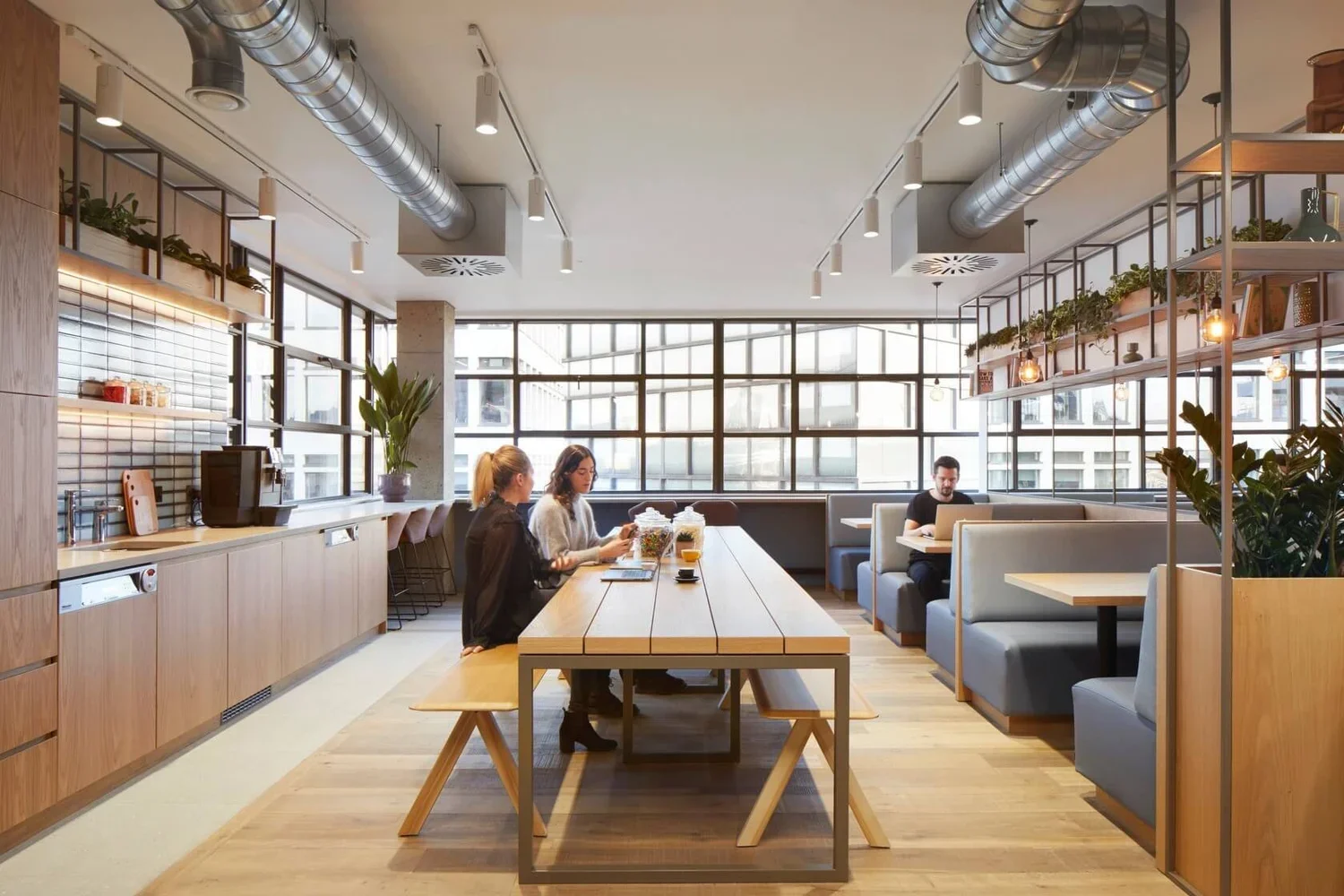 Modern cafe interior with wooden accents, large windows, and a mix of seating options including booth seats and chairs. Three people sit at a long wooden table, working or chatting, with plants and shelves as decor. The space has an industrial ceilin