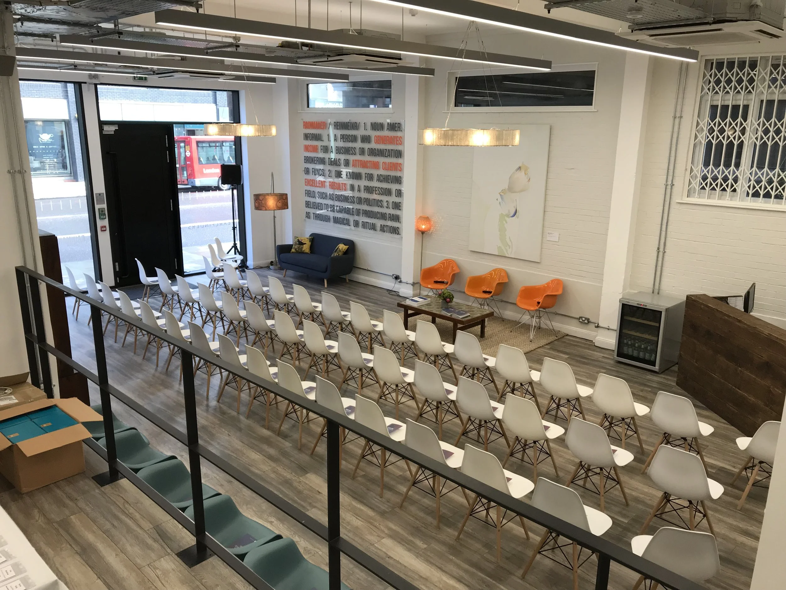 A modern indoor event space with rows of white chairs arranged facing a small stage area. There are two orange chairs, a small table with a decorative plant, and a blue couch with pillows. On the wall, there's a large poster with a definition of the 