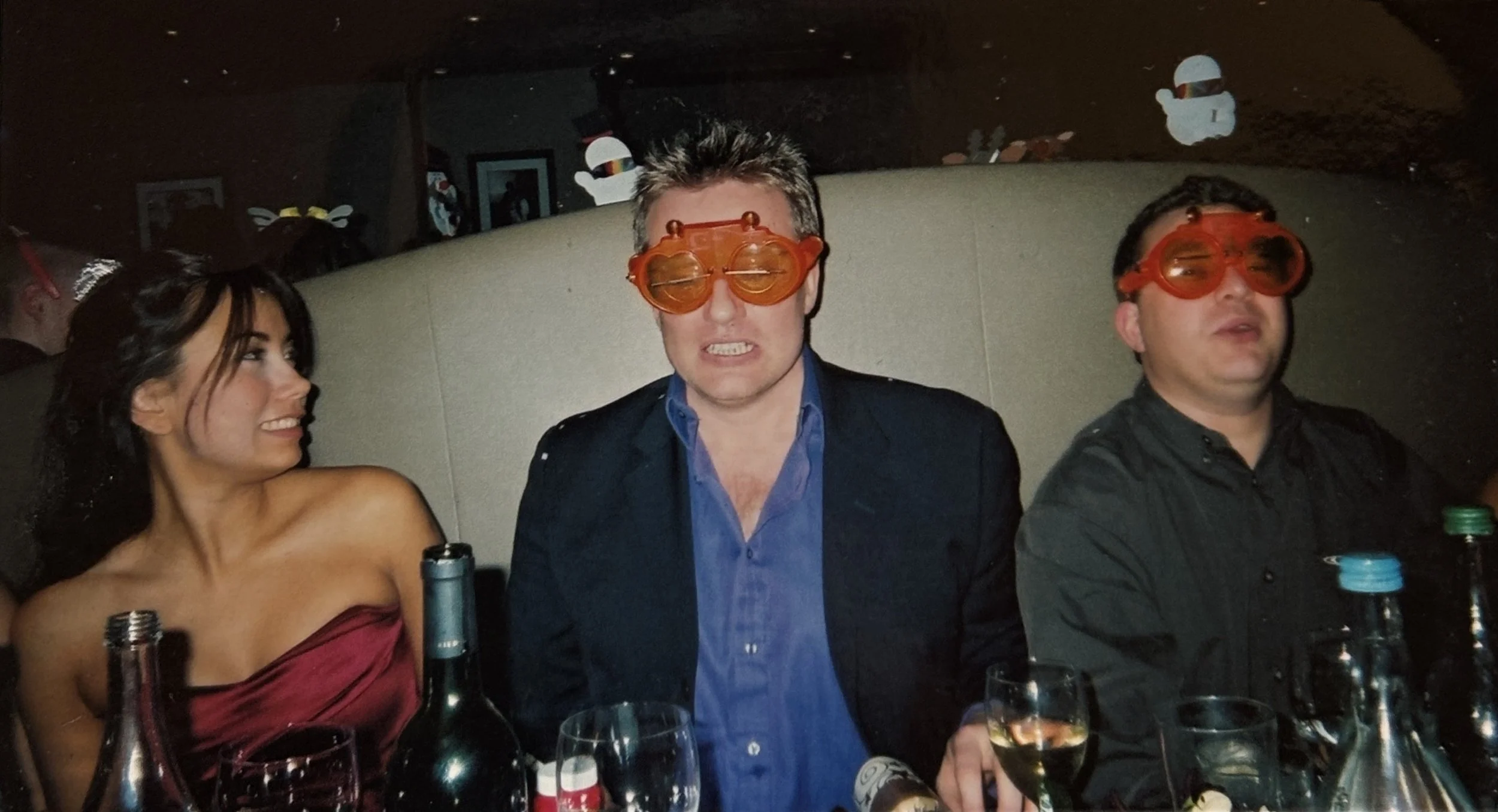 Three people sitting at a table with bottles and glasses, wearing humorous orange glasses, in a dimly lit setting.