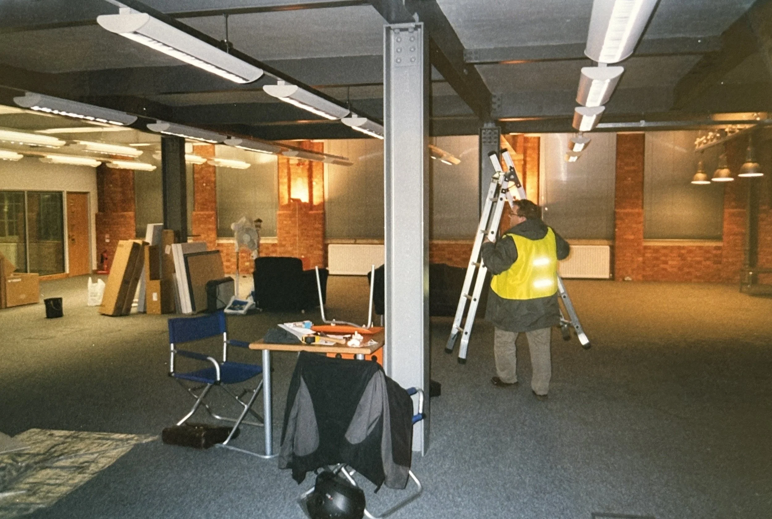 An indoor space with exposed brick walls and modern lighting. A person wearing a yellow safety vest and glasses is standing on a ladder, possibly working on the ceiling. There are chairs, some boxes, and various equipment scattered around the room, i