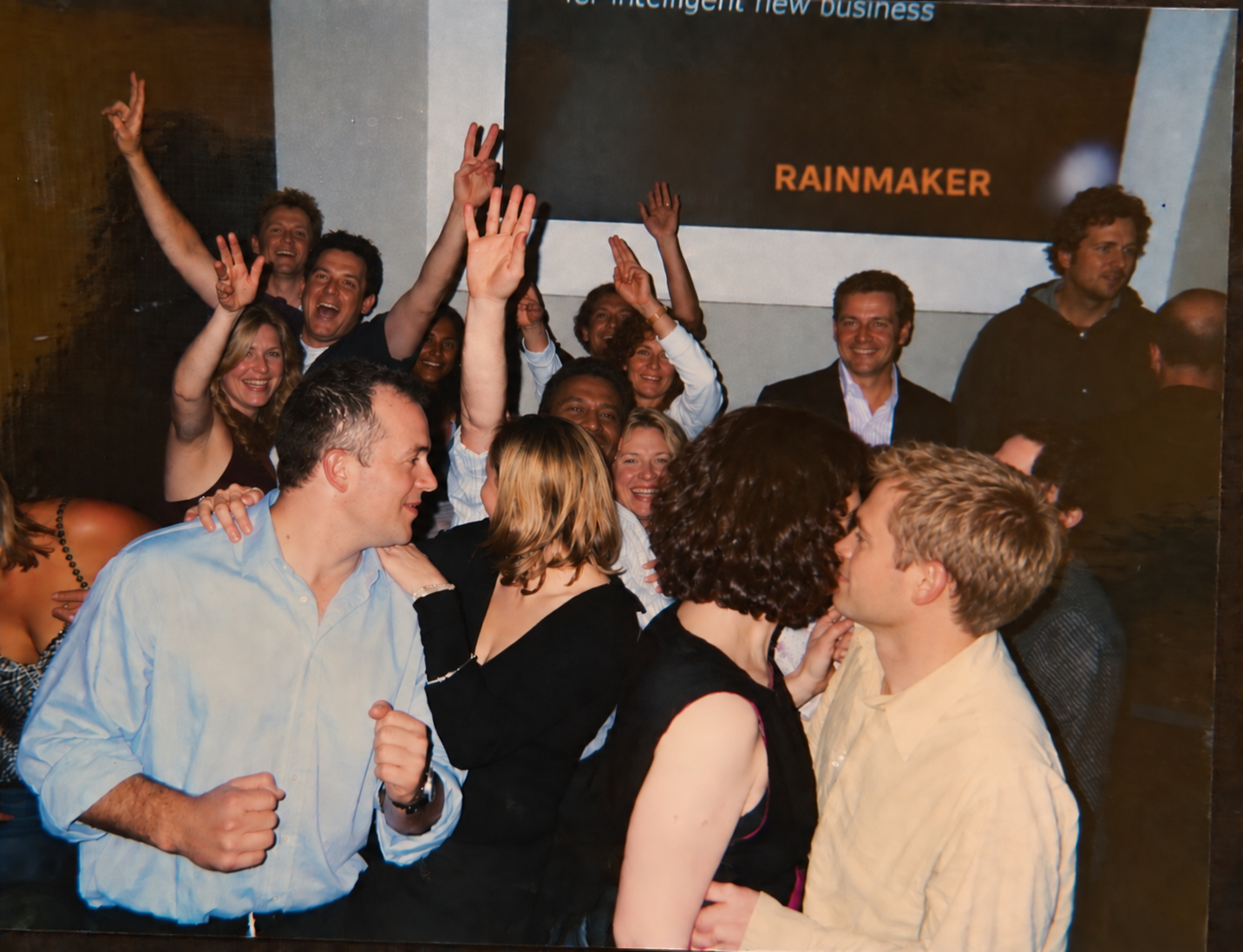 Group of people celebrating at a party, smiling, hugging, and raising their hands. There is a sign in the background with the word 'RAINMAKER'.
