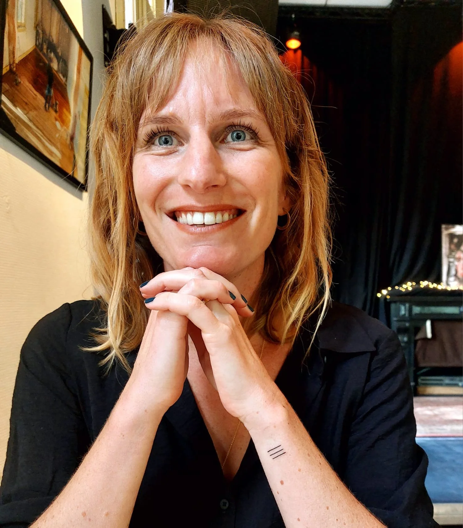 A woman with red hair and blue eyes smiling, sitting with her hands clasped and resting on her chin, in a cozy indoor setting with artwork and warm lighting.