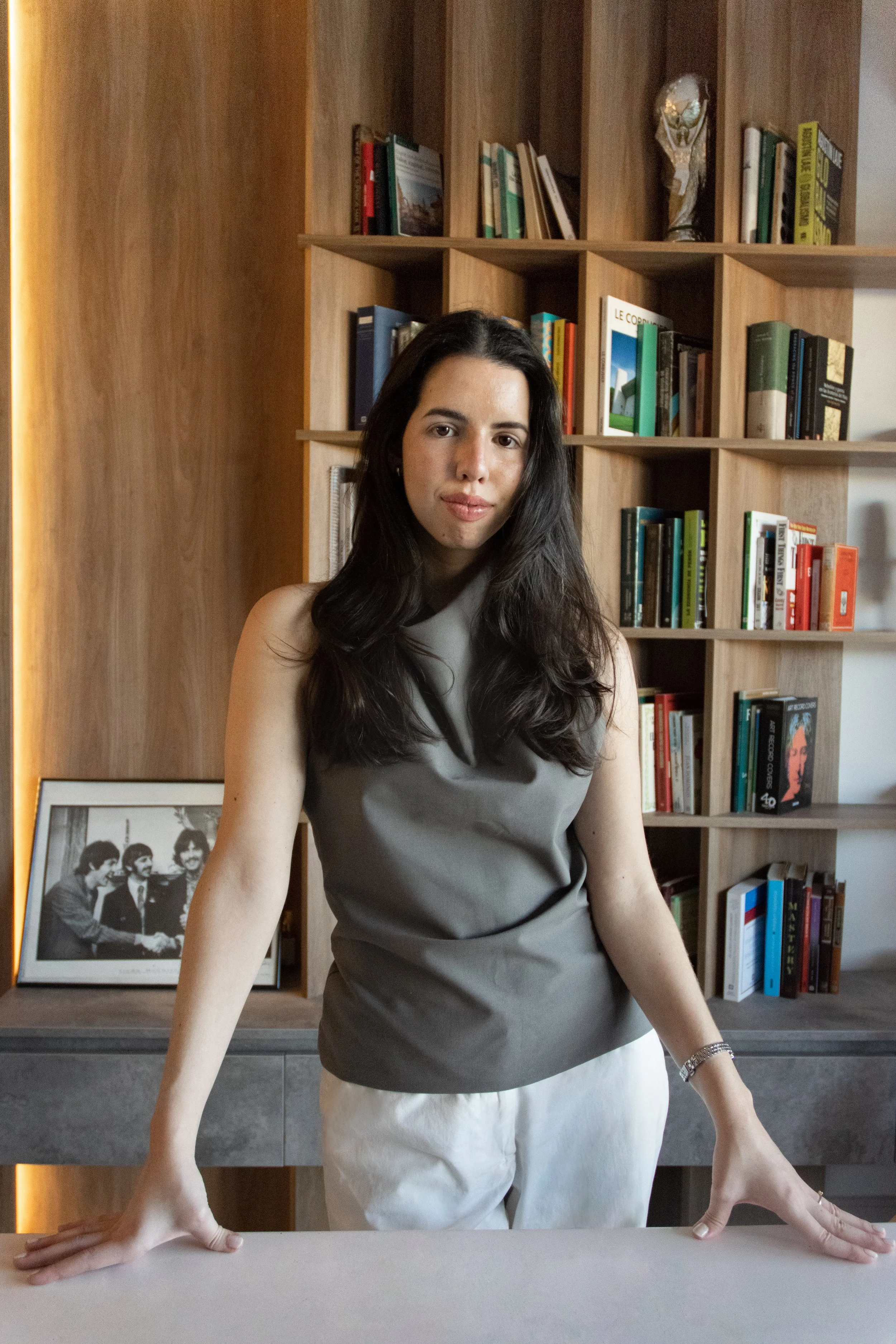 A woman with long dark hair standing behind a table, with her hands resting on the surface. Behind her, there is a wooden bookshelf filled with various books and a black and white framed photo of The Beatles. The setting appears to be indoors, possibly a library or study room.