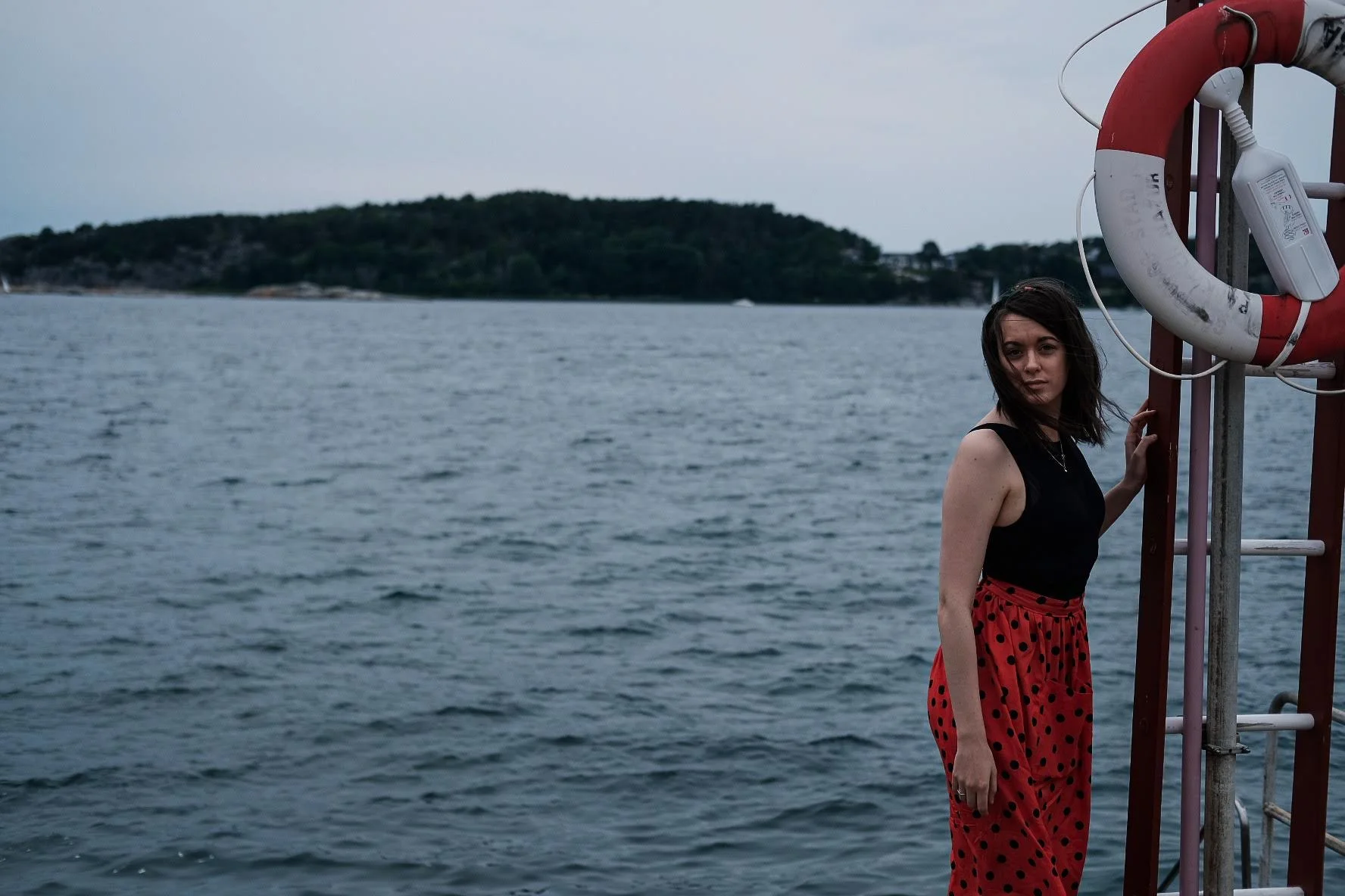 A woman with dark hair wearing a black top and a red polka-dot skirt, with water and an island in the background.