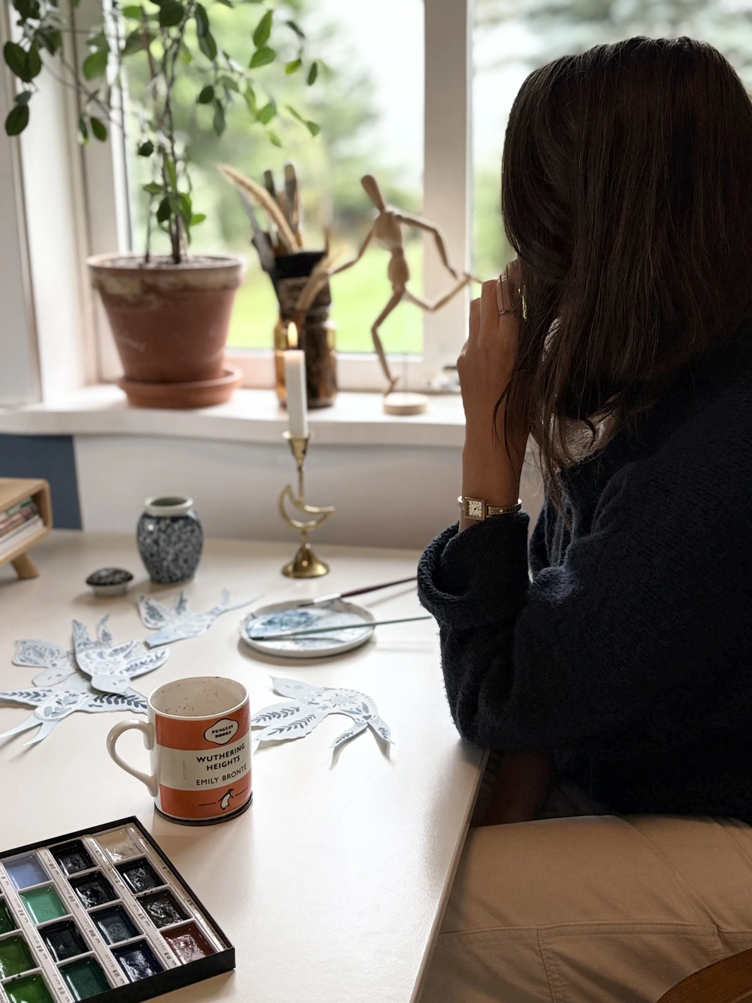 A person with dark hair sitting at a table, holding a camera or phone, looking out a window with potted plants and art supplies on the windowsill. The table has paper cutouts, a mug, and watercolor paint set.