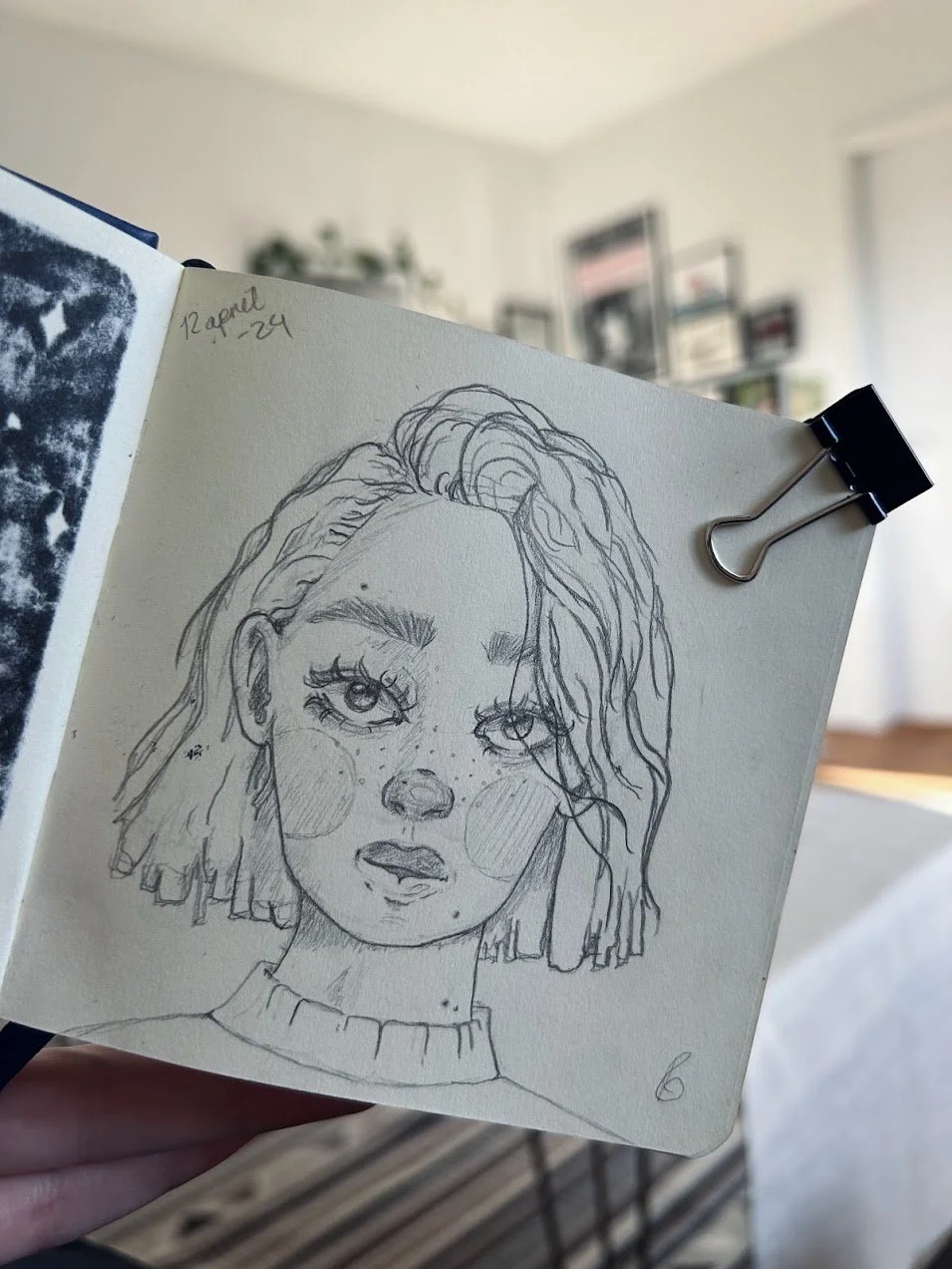 A sketch of a young woman with wavy hair, freckles, large expressive eyes, held in a sketchbook, in a well-lit room with shelves and framed pictures on the wall.