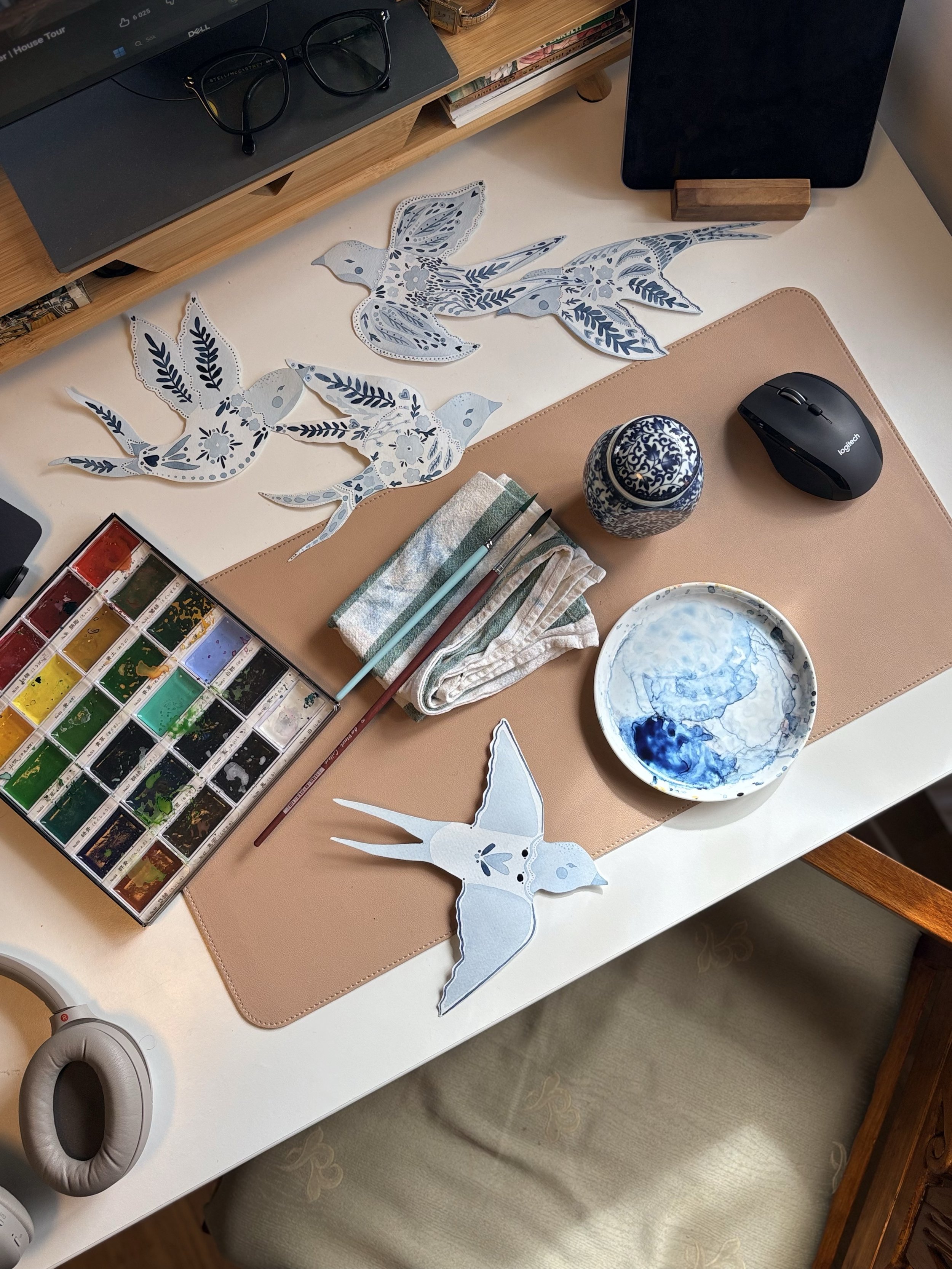 Art supplies on a desk, including watercolor paints, paintbrushes, paper cutouts of birds with blue and white patterns, a small bowl with blue designs, a pair of glasses, computer mouse, and a notebook.