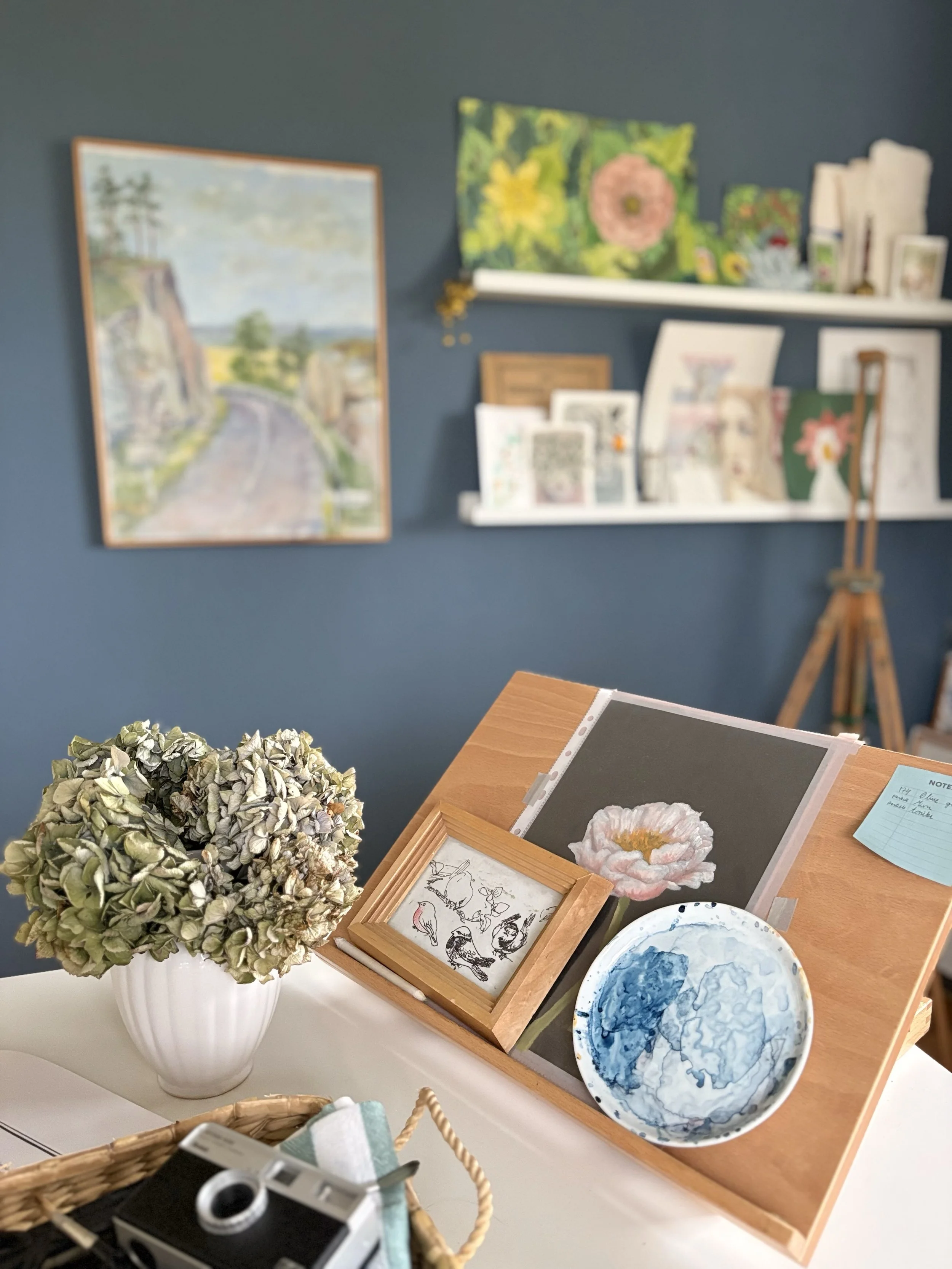 A table with a flower painting, a black and white cityscape, a sketch of birds, with a vase of dried hydrangeas in the foreground, and art prints and a wooden easel in the background.