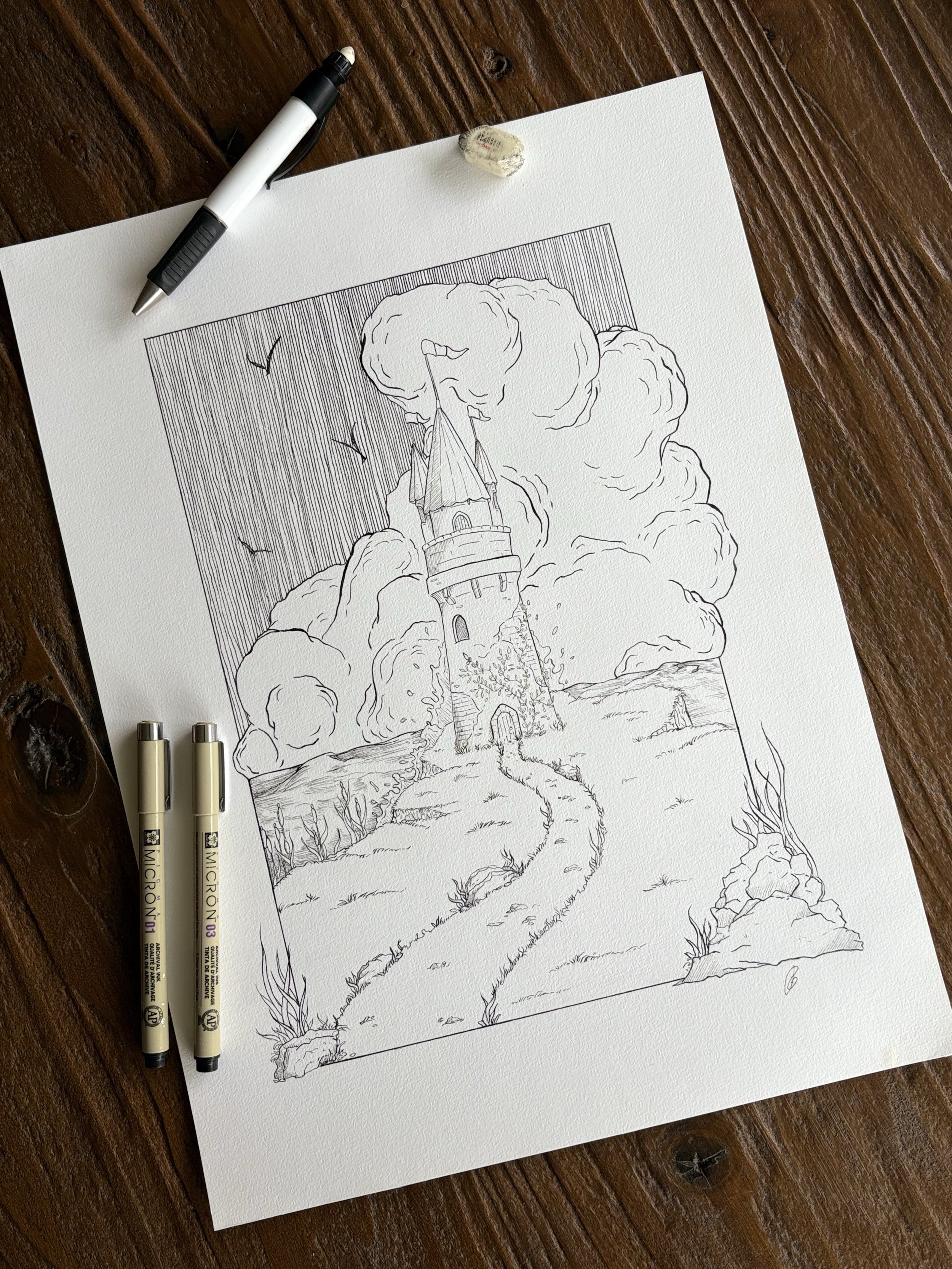 A black ink drawing of a tower with a pointed roof, surrounded by tall clouds and flying in the sky, with a path leading to the castle entrance, on a white textured paper placed on a wooden surface with two markers and an eraser nearby.