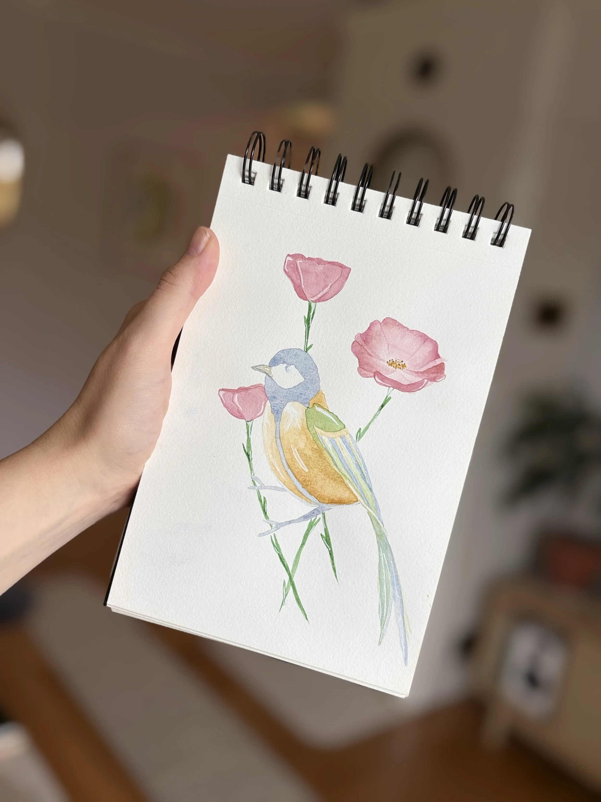 A hand holding a spiral-bound watercolor painting of a bird with pink flowers and green stems on white paper.