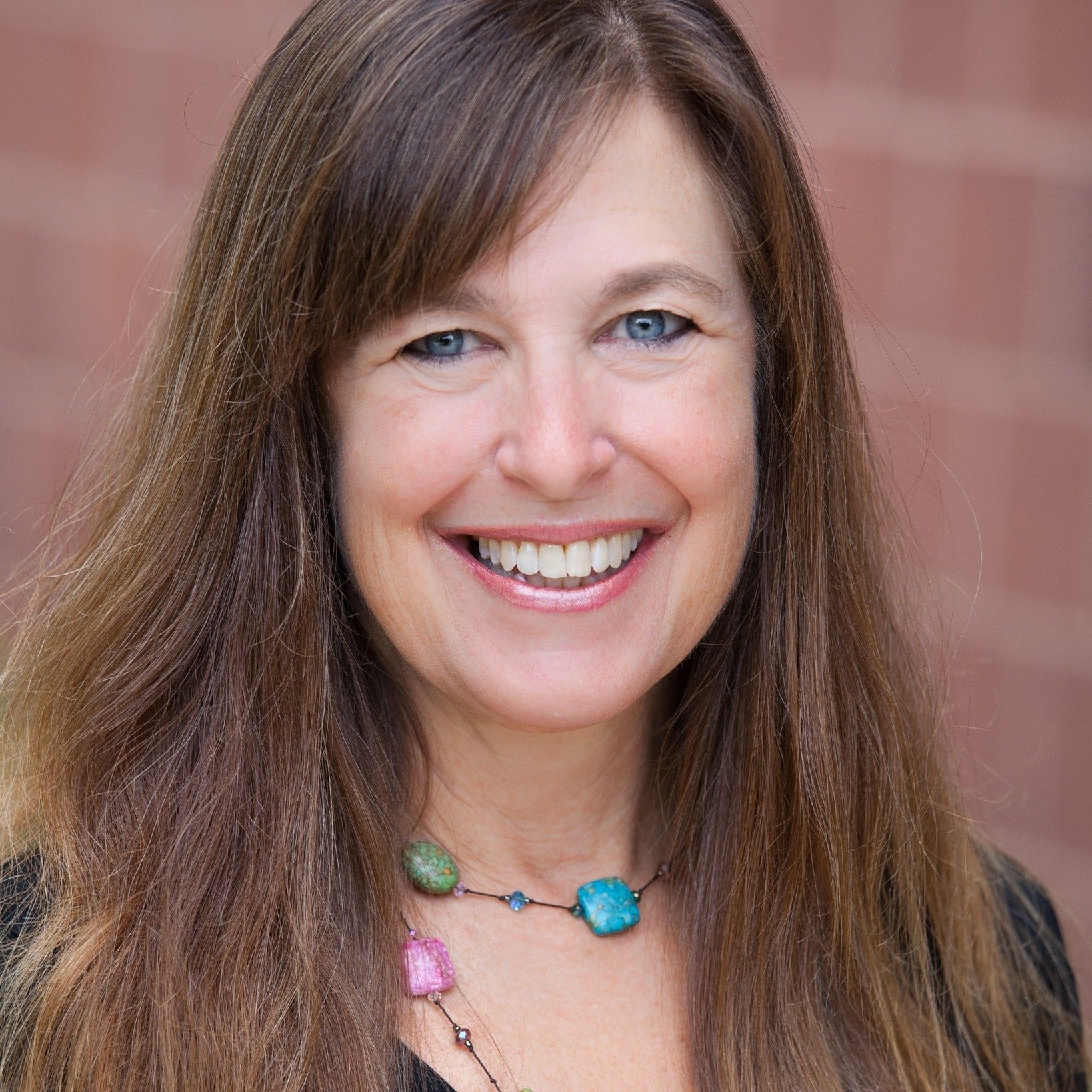 Close-up of a woman with long brown hair, blue eyes, smiling showing her teeth, wearing a colorful beaded necklace, against a blurred pink background.