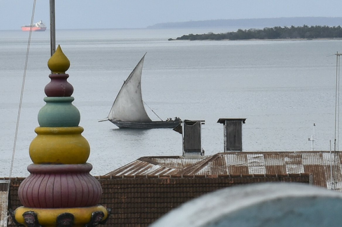 A sailboat with a large white sail on calm water, with a cargo ship in the distance and rural shoreline with trees in the background; foreground includes a colorful, multi-tiered ornamental structure and rooftops.