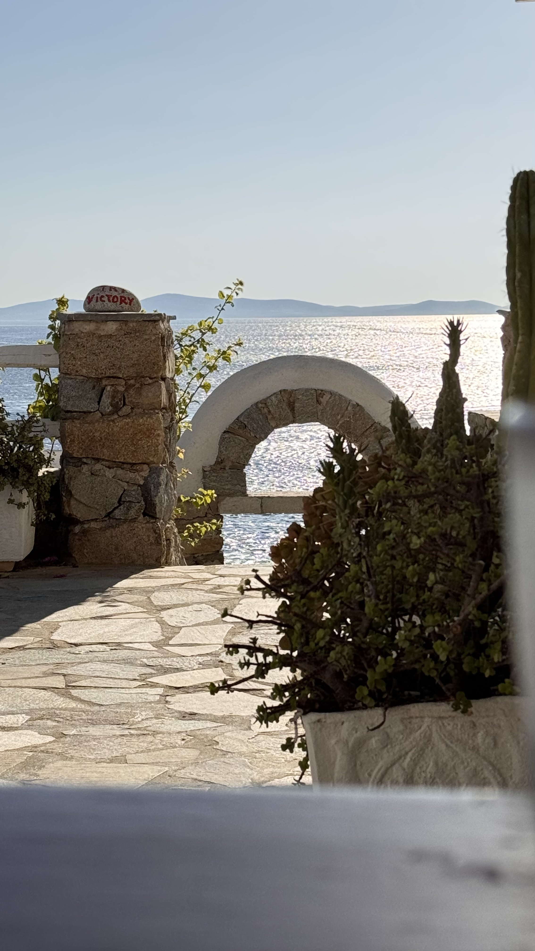 A scenic view of a body of water with distant mountains, seen through a stone archway and a brick pillar with a painted rock that says 'Victory,' surrounded by plants and outdoor decor.