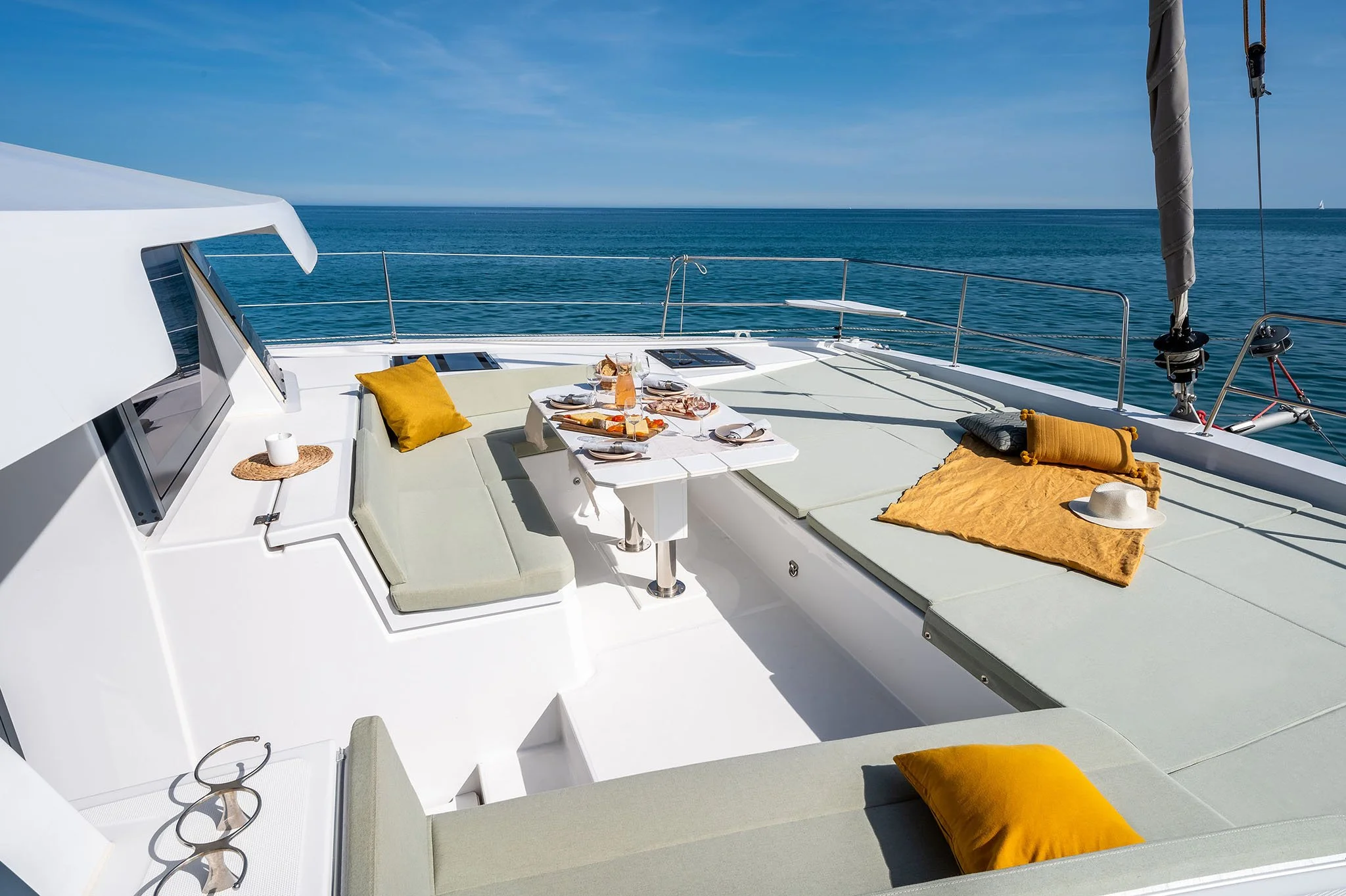 A yacht with a dining table set for a meal, surrounded by cushions, on the deck overlooking the ocean under a clear blue sky.