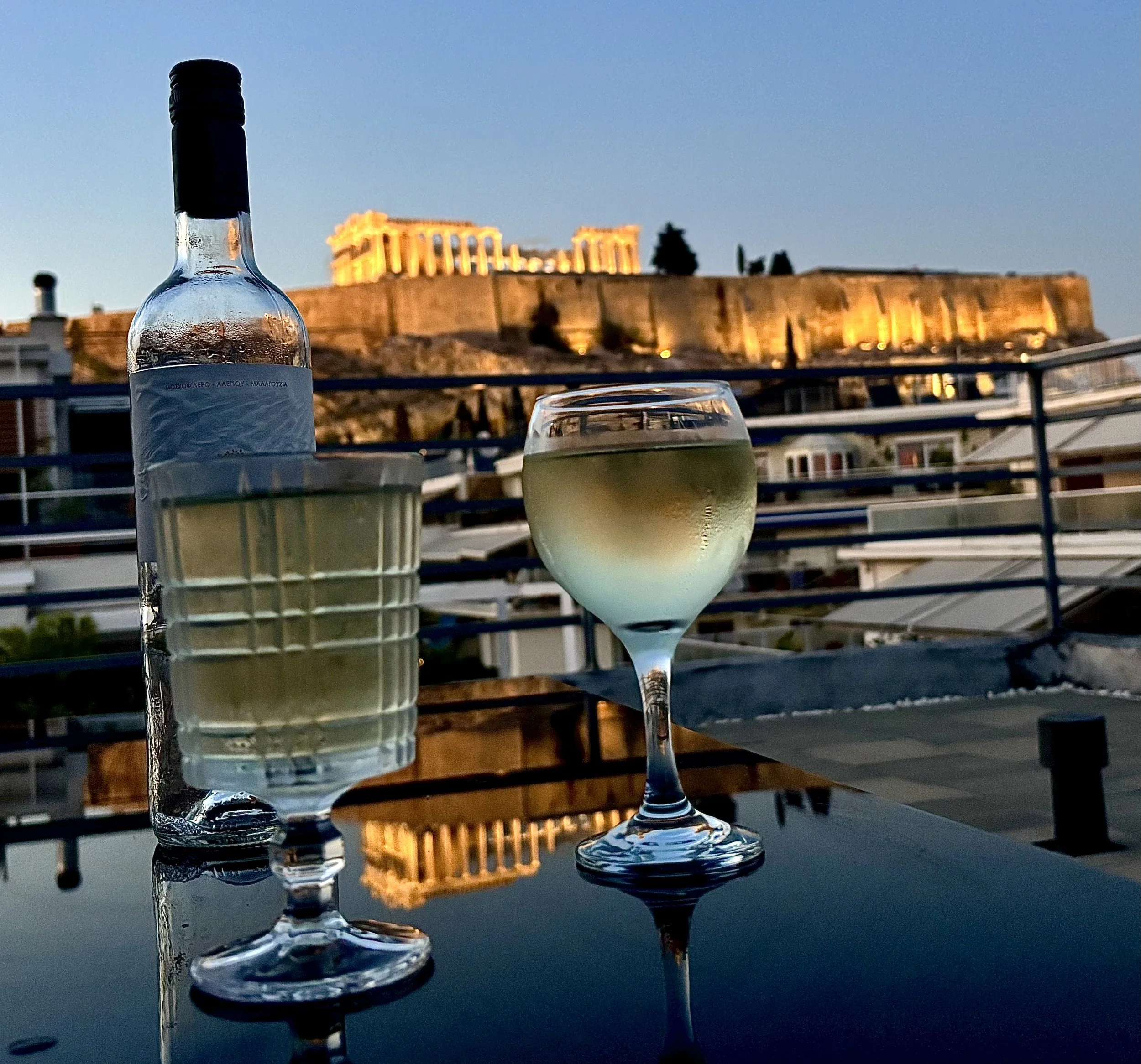 A bottle of water and two glasses of white wine on a reflective surface with a view of the illuminated Parthenon atop the Acropolis in Athens, Greece, during dusk.