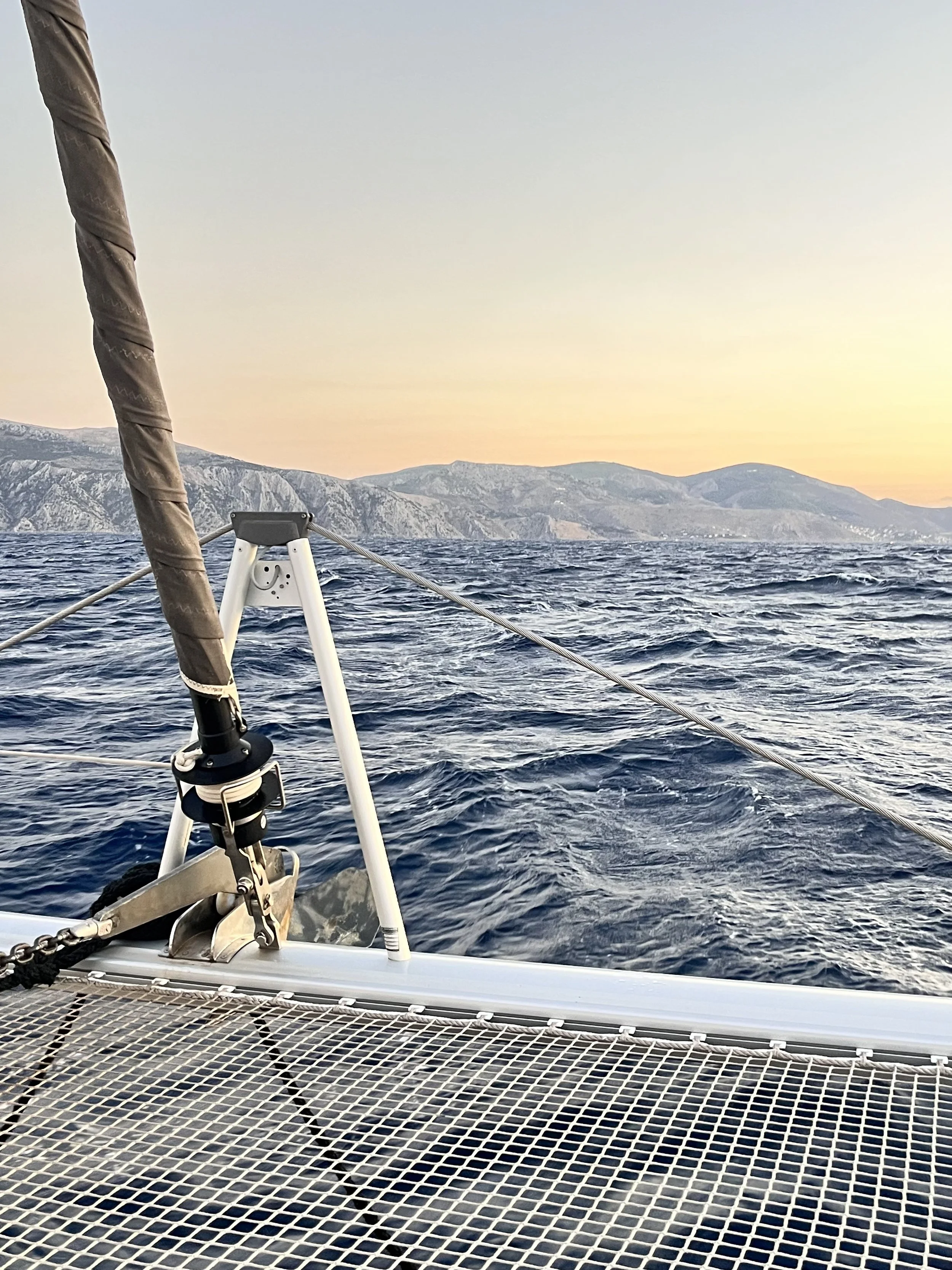View from a boat showing a metal grid deck, rigging, and part of the boat's structure in the foreground, with choppy ocean water and distant landforms or mountains at sunset in the background.