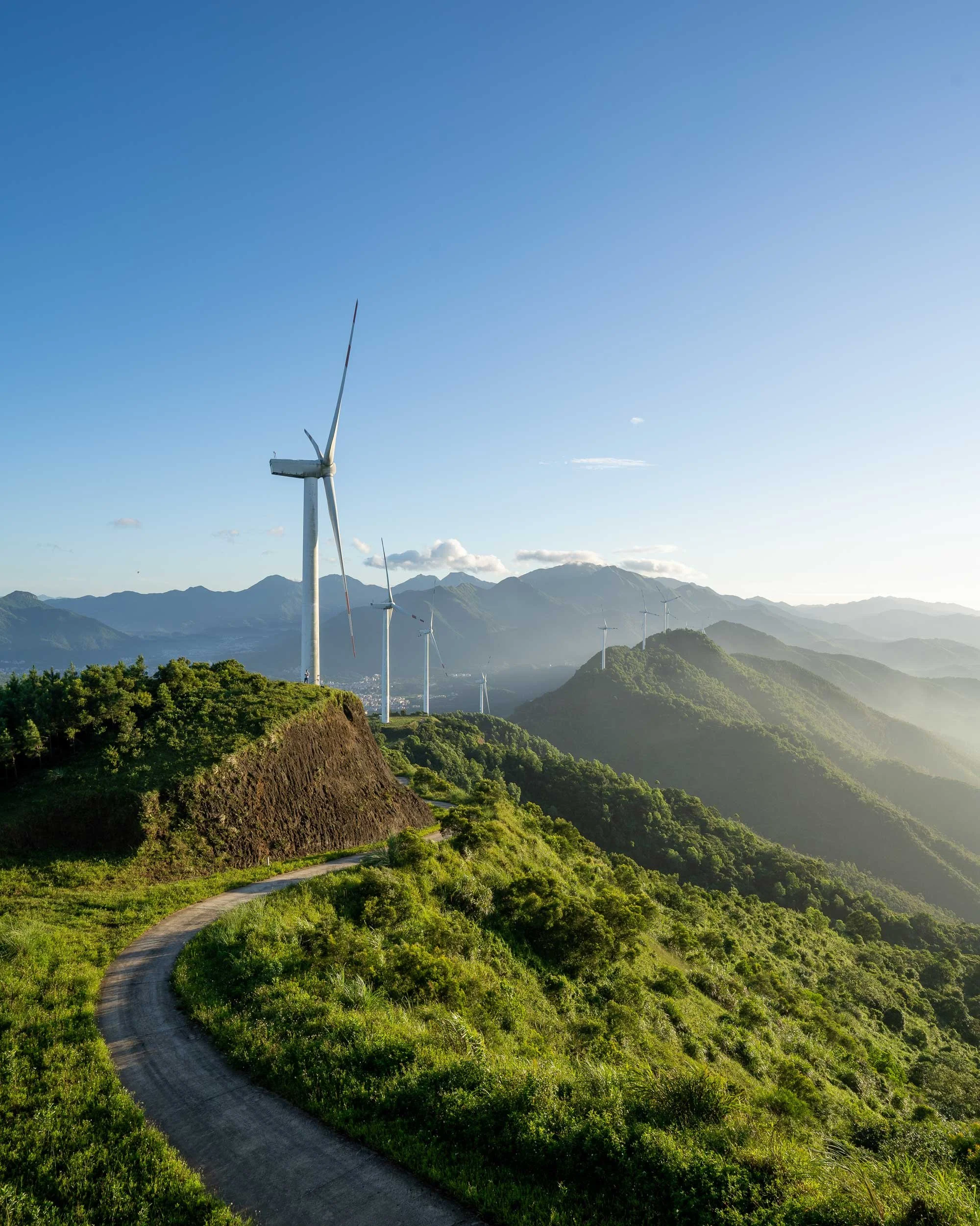 Wind turbines on a green hillside with a winding dirt road, mountains in the background, and a clear blue sky.