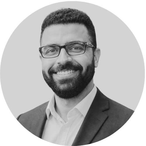 Black and white headshot of a man with glasses, a beard, and short hair, wearing a suit jacket and white shirt, smiling.