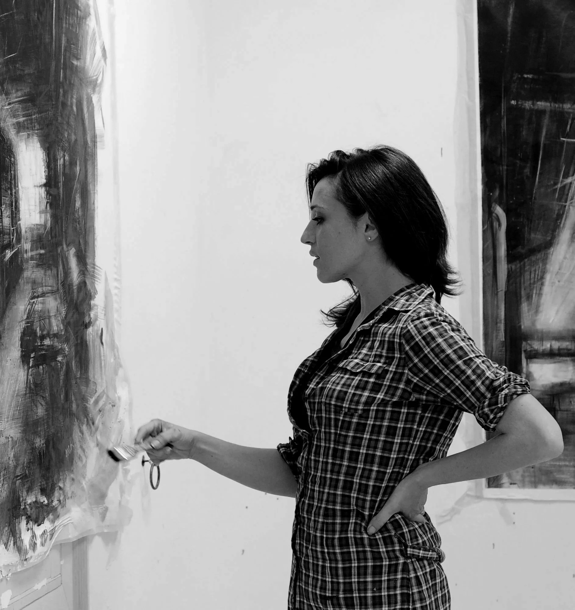 A woman with dark hair wearing a plaid shirt, standing sideways, looking at a canvas in an art studio.