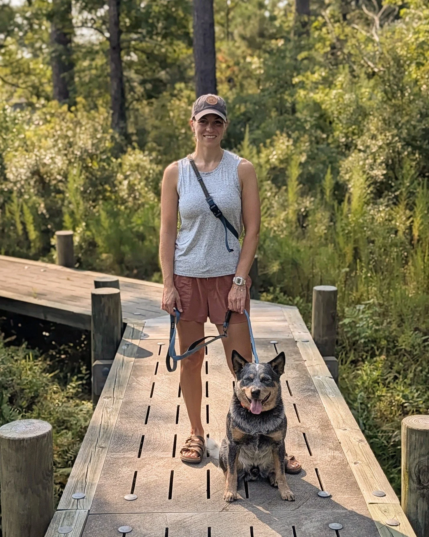 A woman standing on a wooden bridge outdoors holding the leash of a dog, with a forest in the background. The woman is smiling, wearing a gray tank top, shorts, sandals, and a baseball cap. The dog is sitting on the bridge, facing forward with its tongue out.