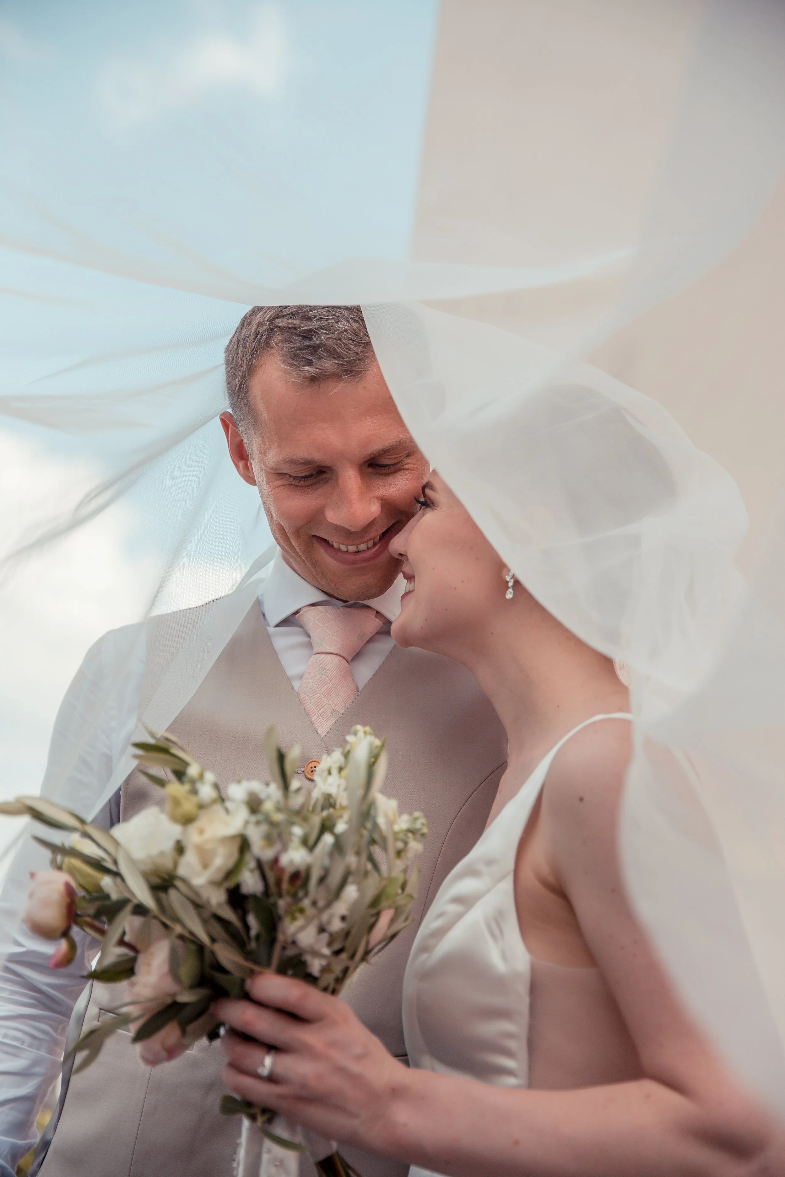 A smiling groom and bride close-up, partially covered by wedding veil, holding a bouquet of flowers.
