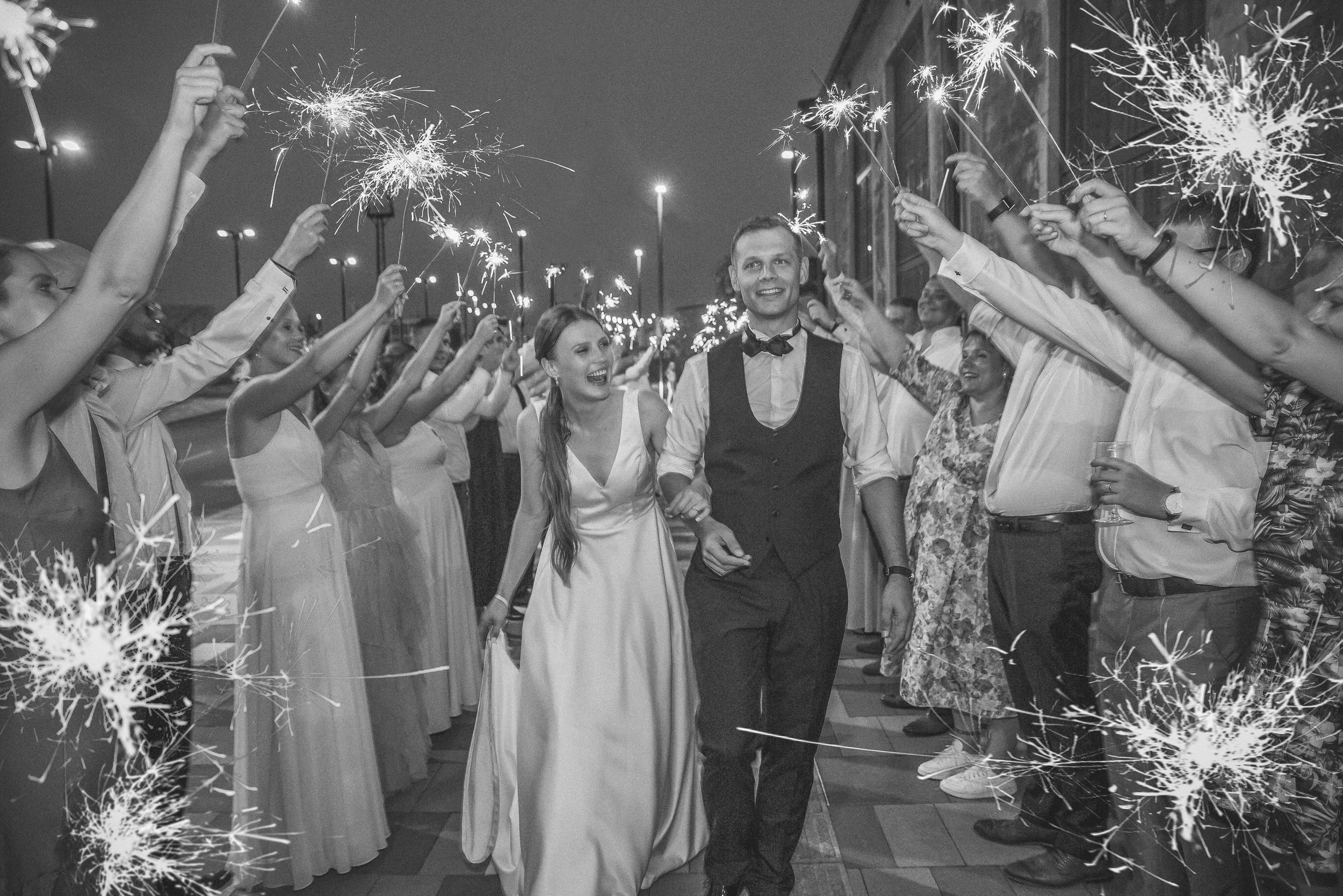 A black and white photo of a happy bride and groom holding hands while walking through a crowd of cheerful guests at night. The guests are holding sparklers and celebrating, with some smiling and looking at the couple.