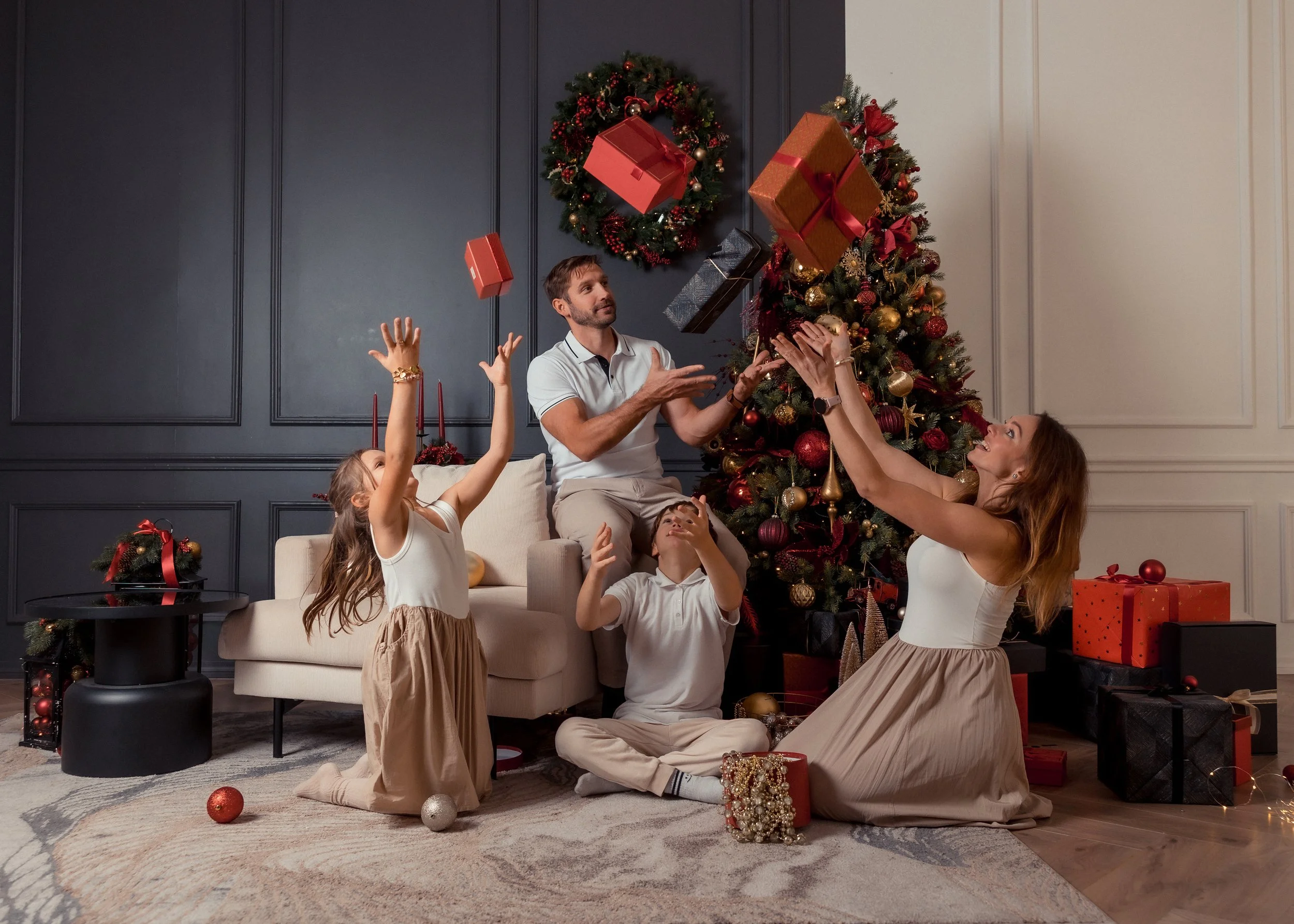 A family of five kids and two adults celebrating Christmas in front of a decorated Christmas tree with gift boxes. The children are reaching to catch falling gifts, and everyone looks joyful.