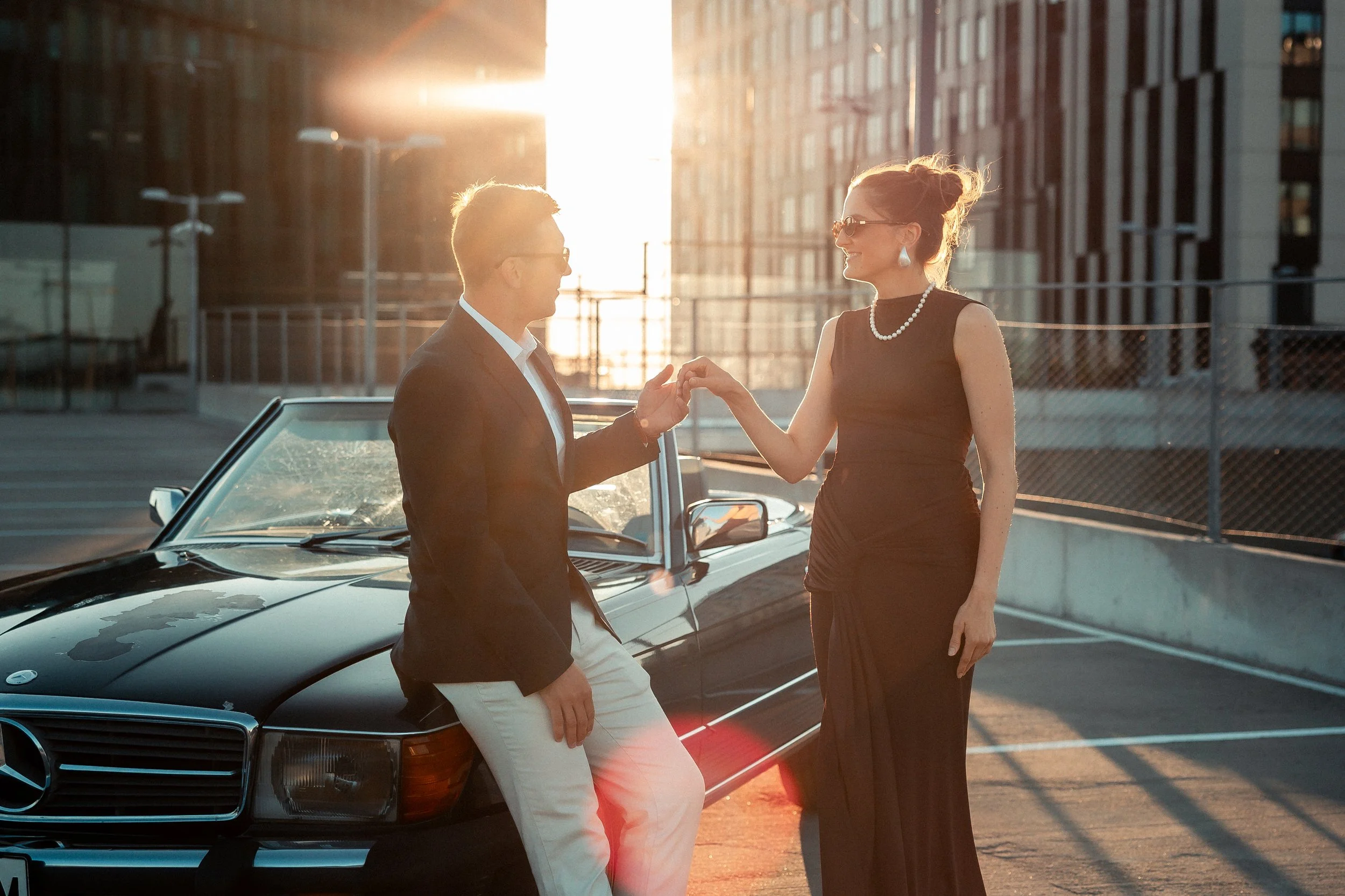 A man in a dark blazer and white pants and a woman in a black dress and pearls exchanging a handshake or touching fingers next to a black luxury car in a parking lot during sunset.