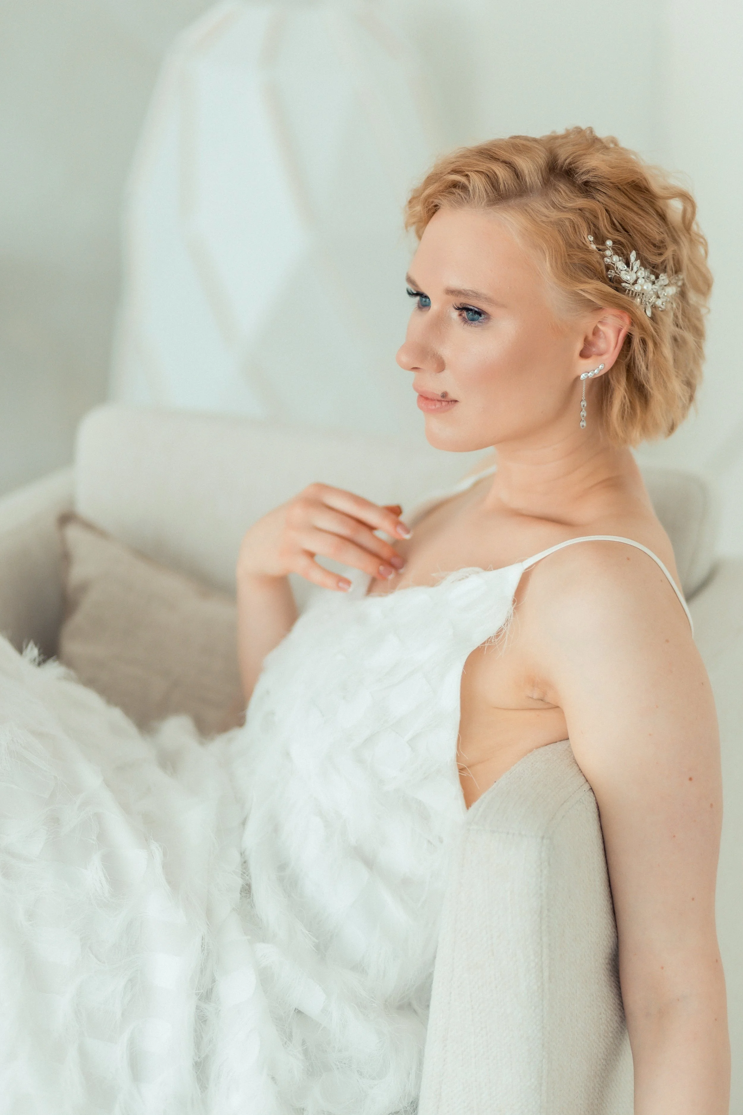A woman with blonde hair styled in soft waves, wearing pearl earrings and a pearl hair accessory, dressed in a white dress, sitting on a beige couch with a white fluffy blanket, in a softly lit room.