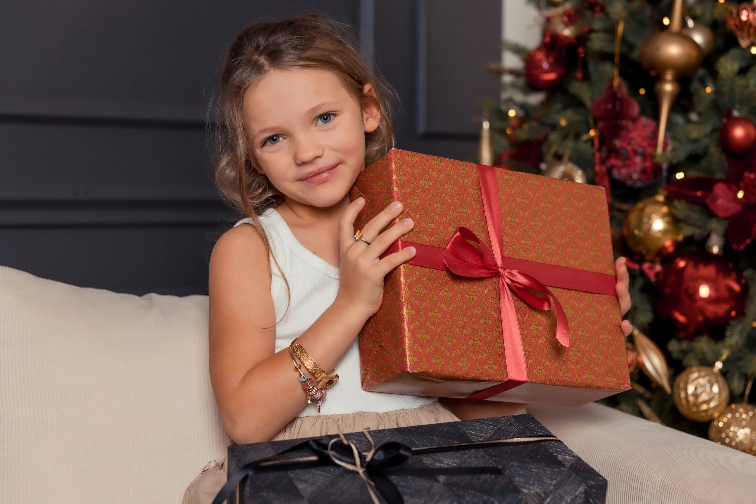 A young girl with blue eyes holding a wrapped Christmas gift in front of a decorated Christmas tree.