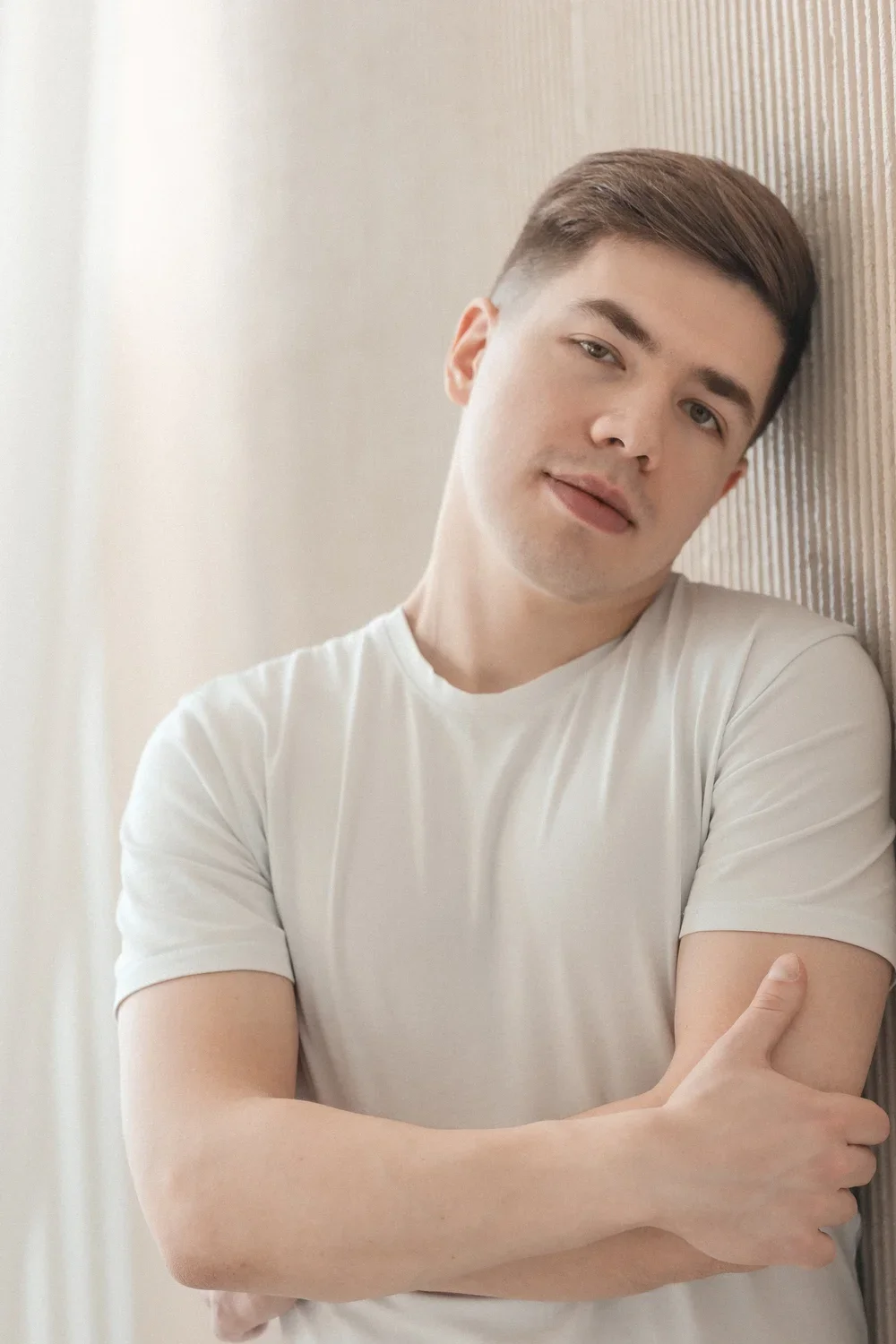 A young man with short brown hair, wearing a white t-shirt, leaning against a wall and looking at the camera with a relaxed expression.