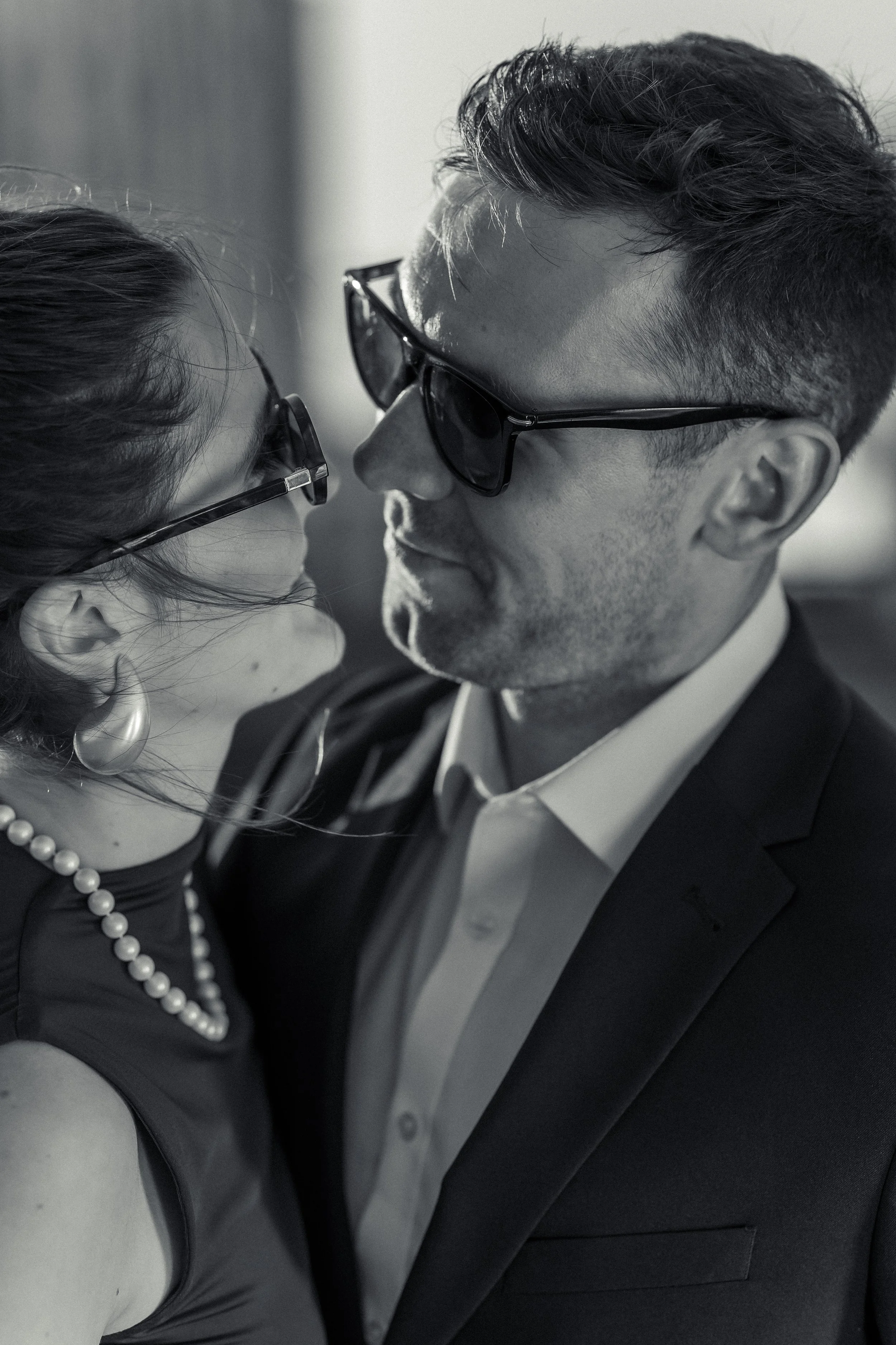 A close-up black and white photograph of a man and woman face-to-face, both wearing dark sunglasses, dressed in formal attire with pearl jewelry, leaning in towards each other.