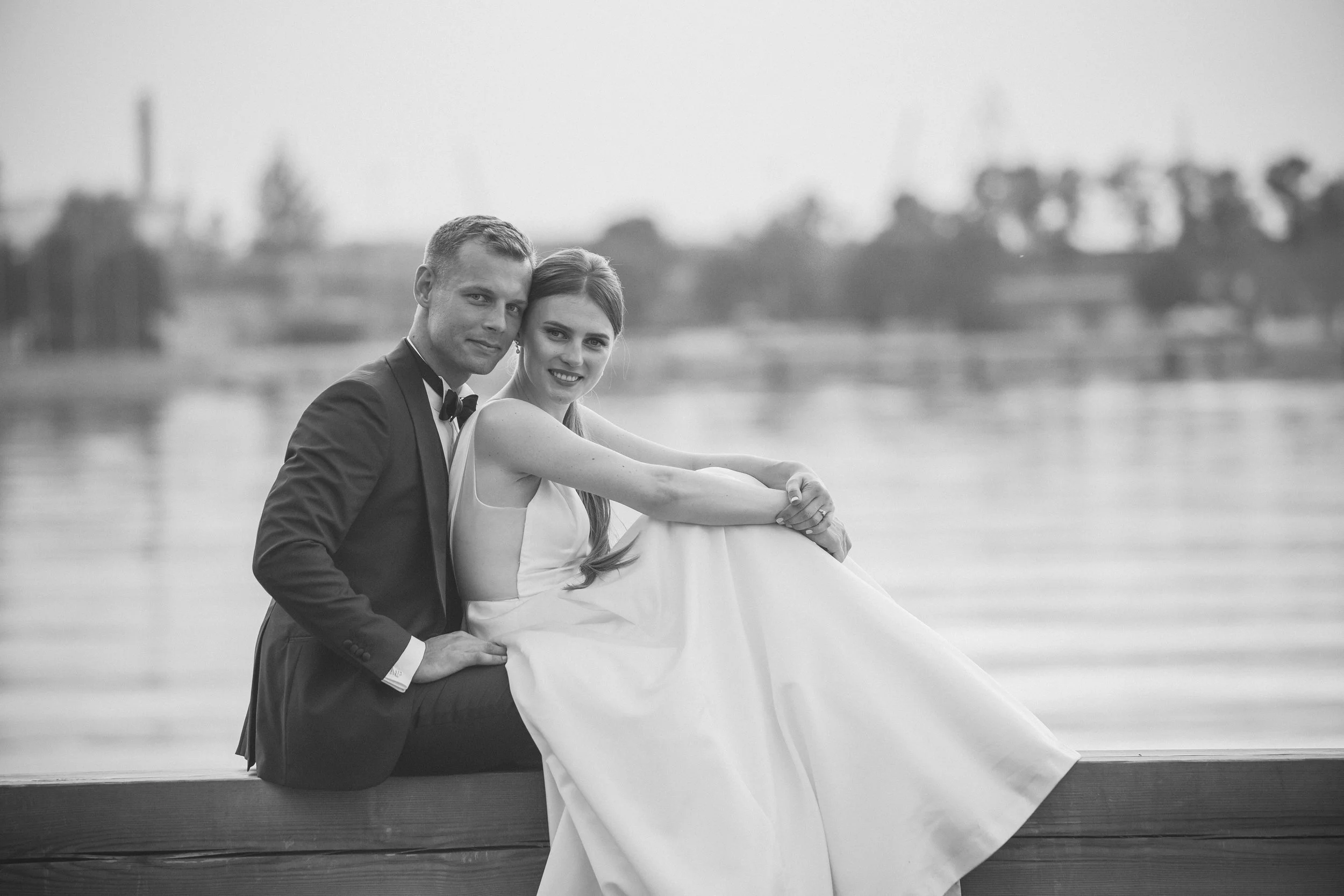 Black and white photo of a newlywed couple, sitting on a bench near water, smiling and embracing each other, with trees and a body of water in the background.