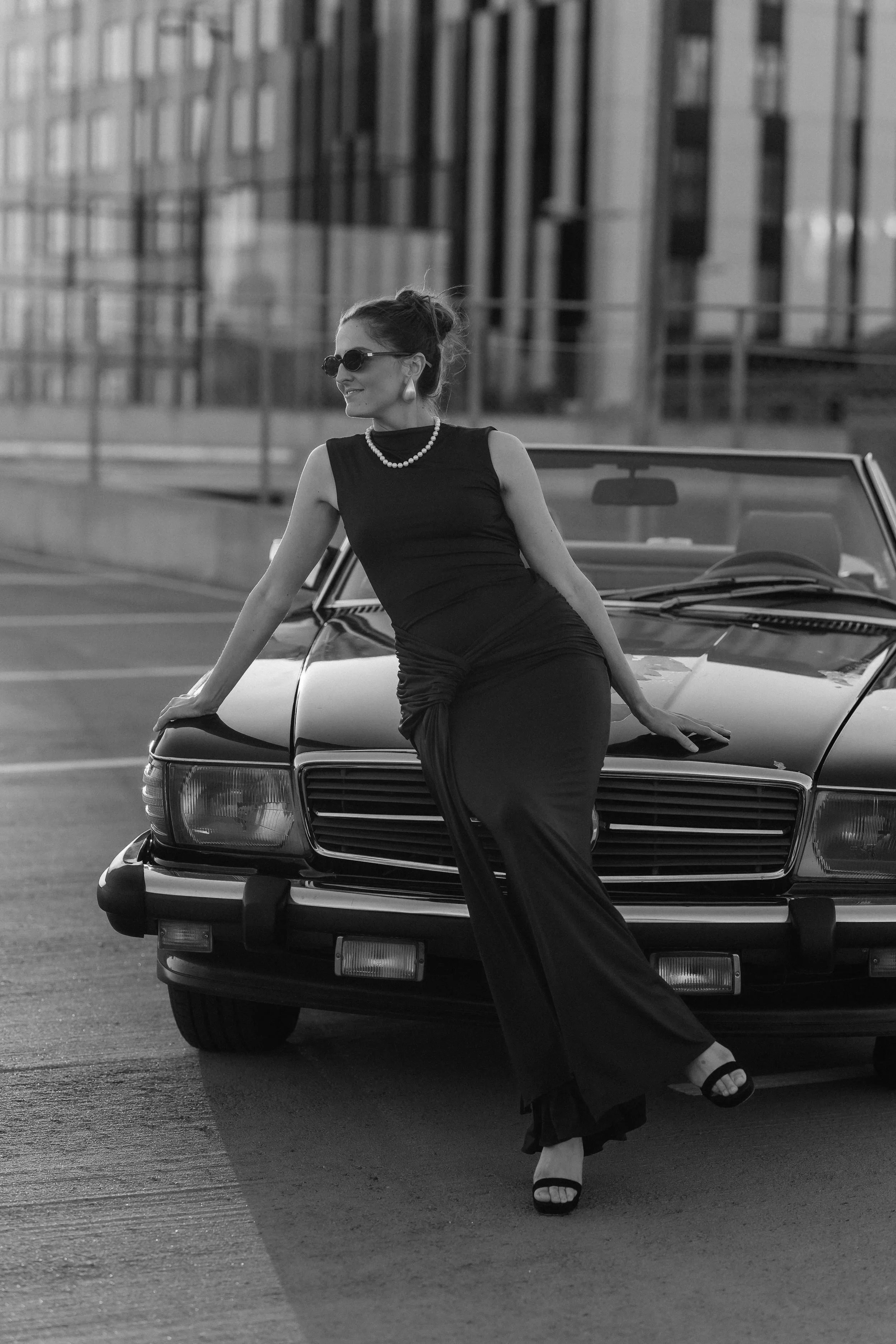 A woman in a black sleeveless dress with pearl necklace and earrings and sunglasses is sitting on the hood of a vintage car with a cityscape background.