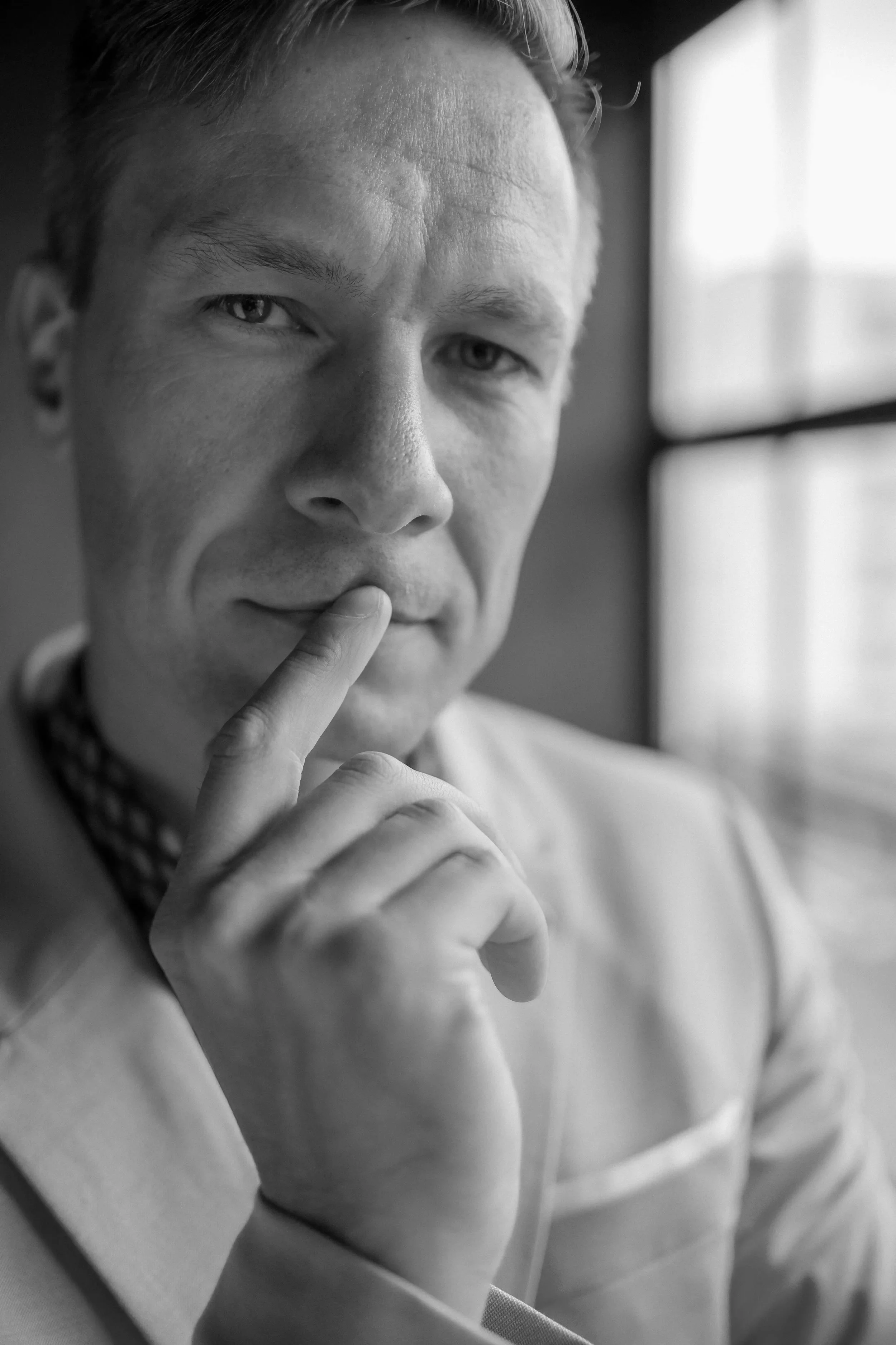 A black-and-white close-up photograph of a person with light skin, short hair, and a thoughtful expression, touching their lips with their fingers while looking out a window.