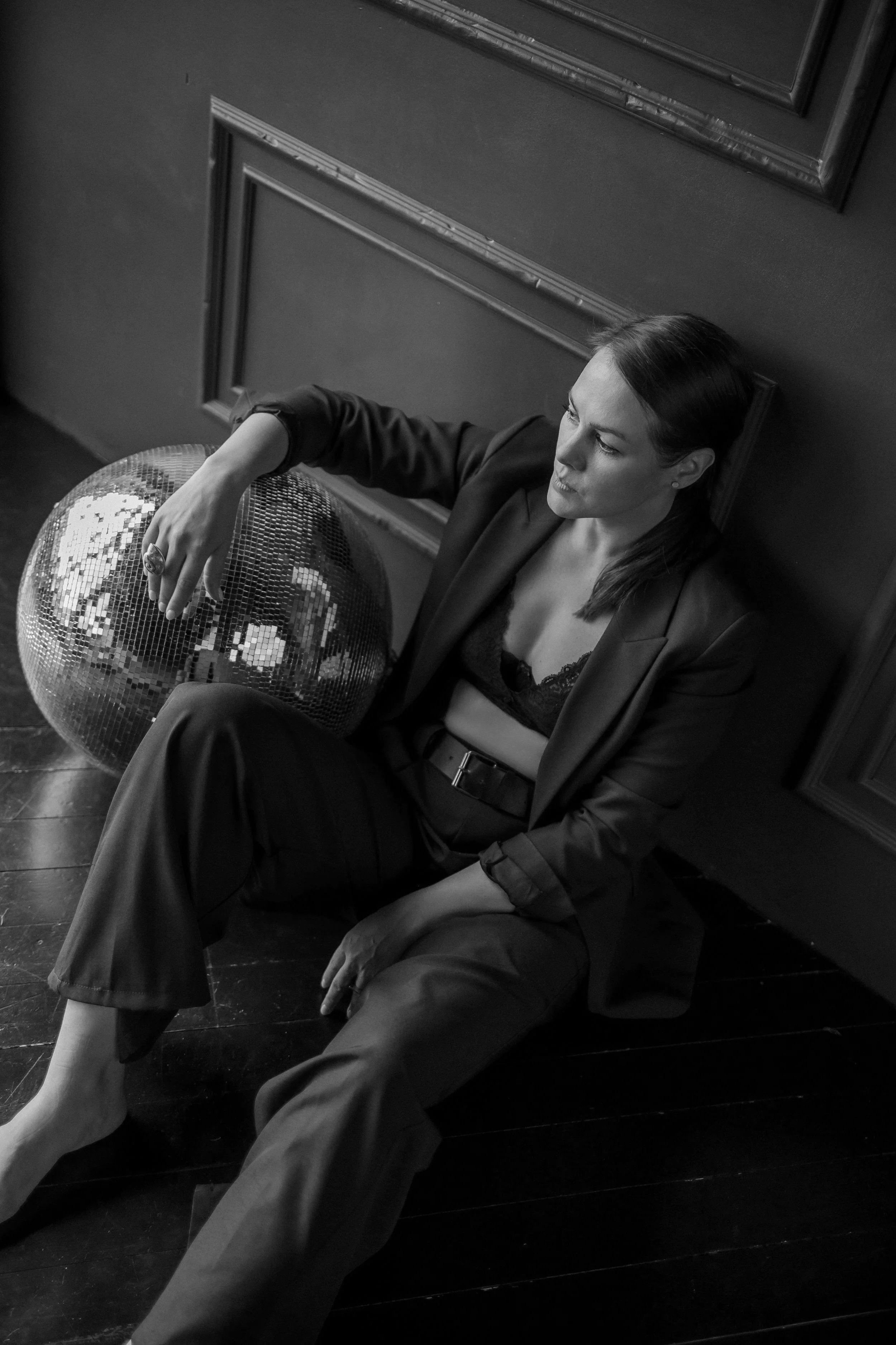 A black and white photo of a woman with dark hair wearing a blazer and lace top, sitting on the floor with a reflective disco ball on her lap, in a room with dark wooden flooring and dark walls with decorative trim.