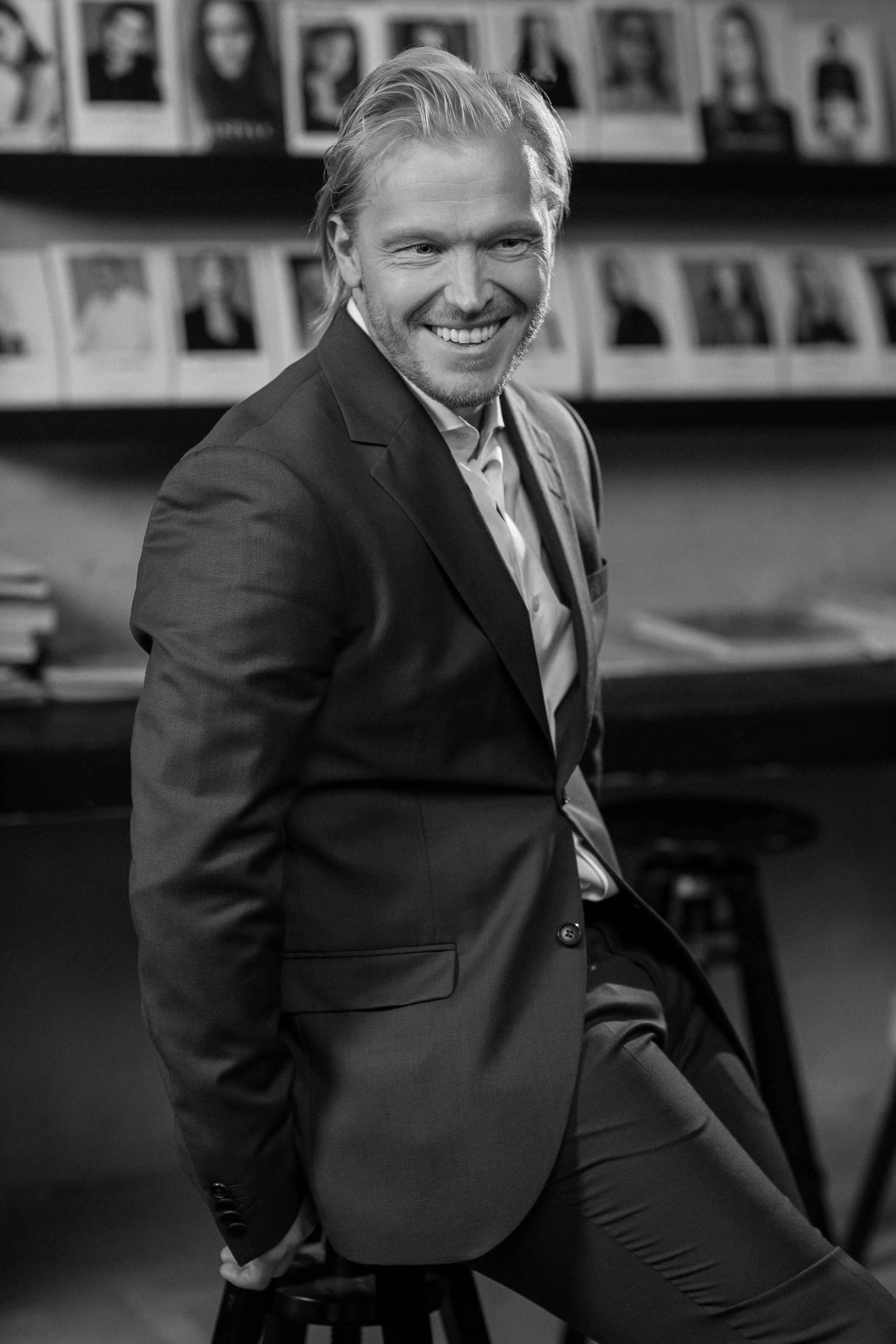 Black and white photograph of a man in a suit smiling, with a background of photos on a wall.
