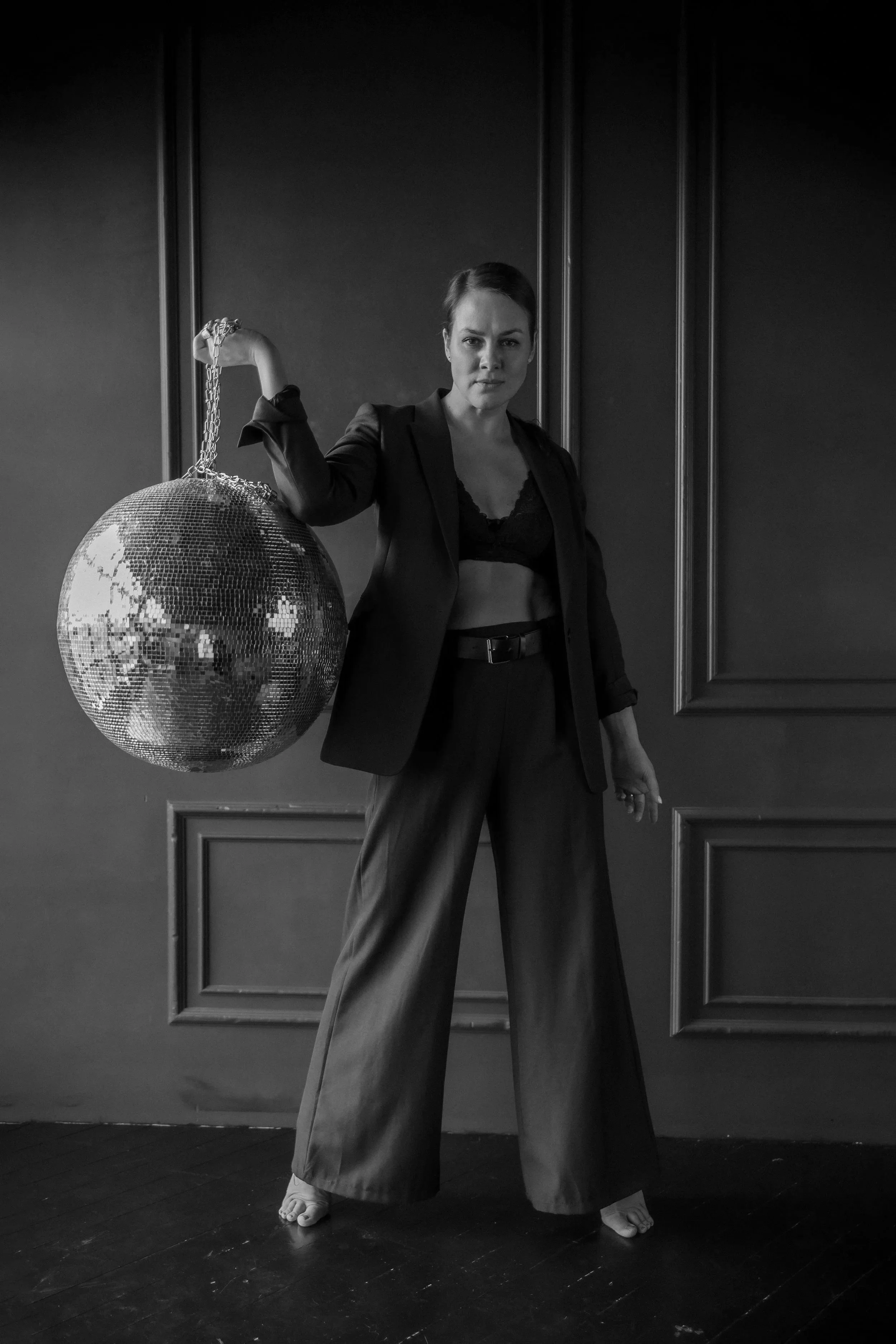 A woman in a black blazer and wide-legged trousers holds a disco ball with a chain in her right hand, standing in a room with wood floors and dark panel walls.