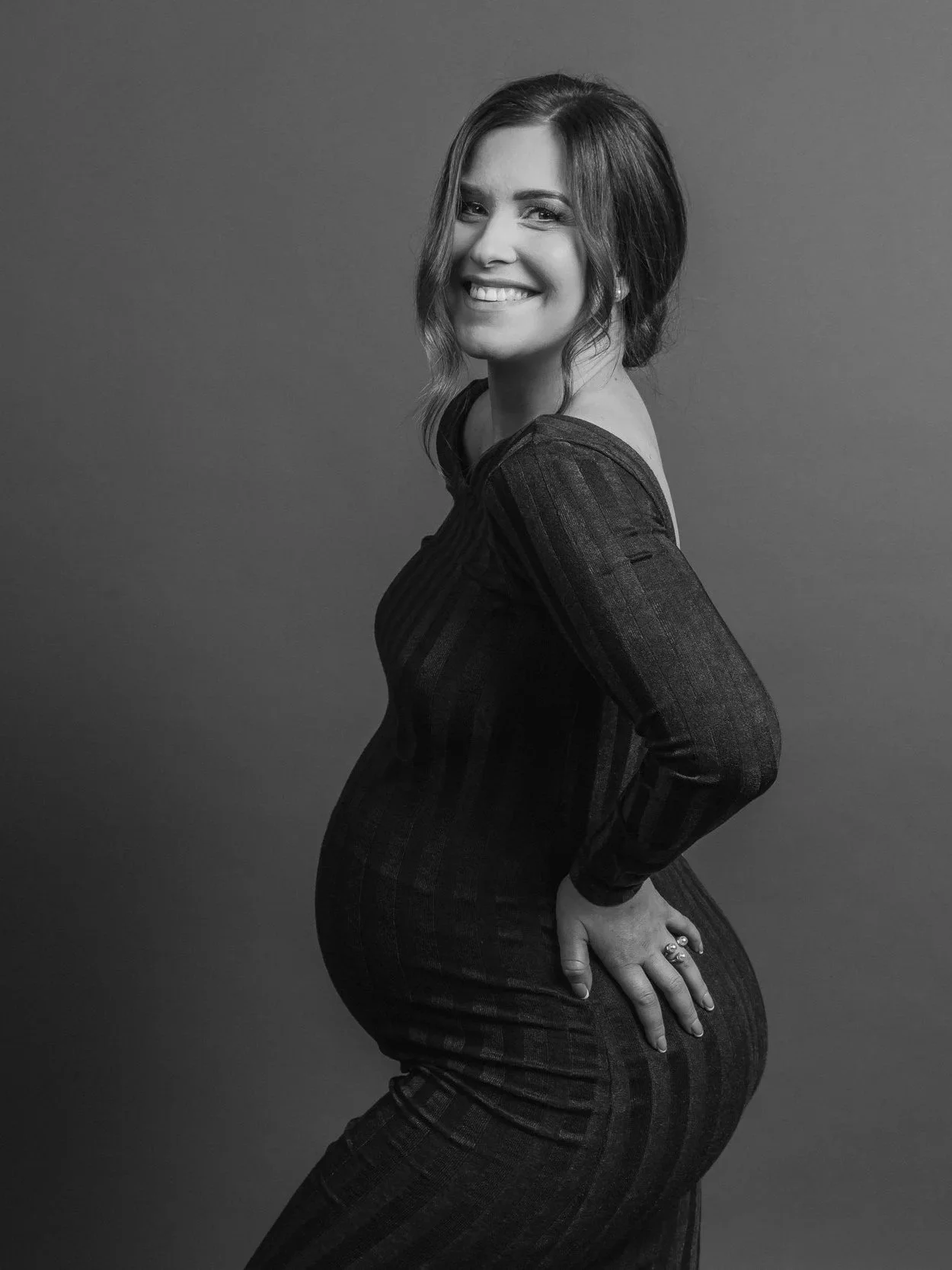 A pregnant woman with shoulder-length hair, smiling, wearing a dark patterned dress, standing sideways with one hand on her hip, in front of a plain wall.