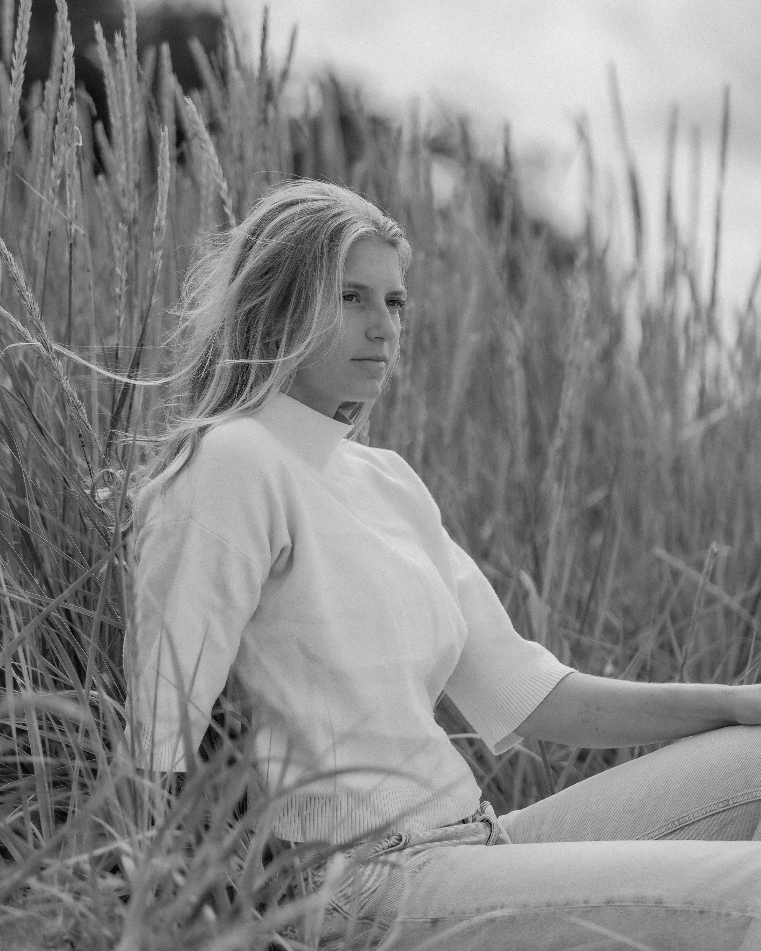 Black and white photo of a young woman sitting in tall grass near a body of water, looking into the distance.