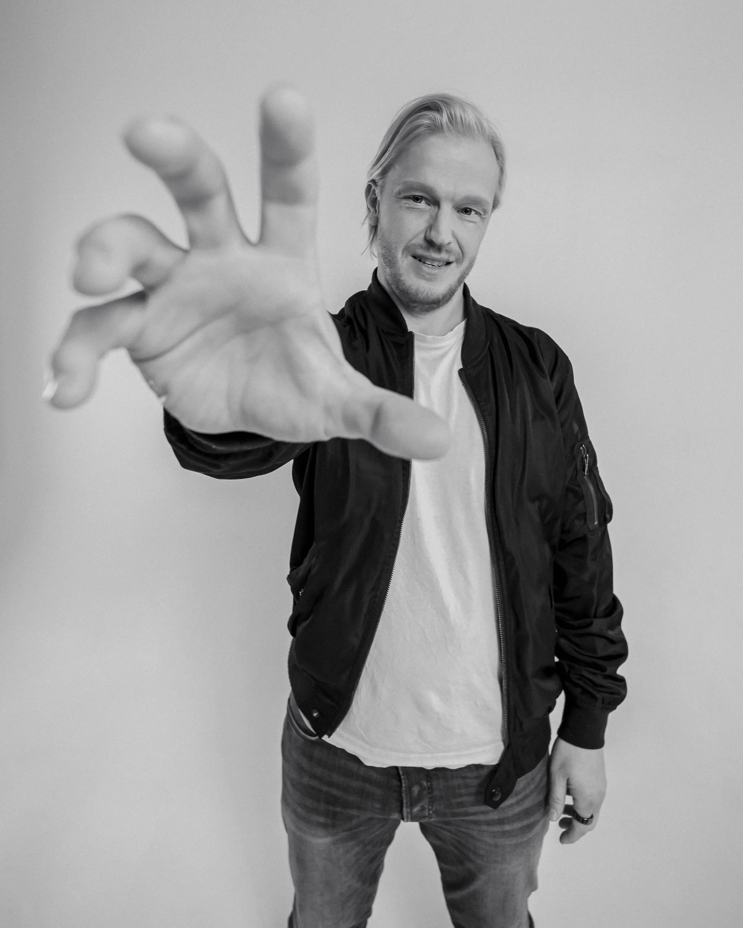 A black and white photo of a young man with blonde hair, wearing a black jacket and gray pants, standing in front of a plain wall. He is smiling and reaching towards the camera with his right hand.