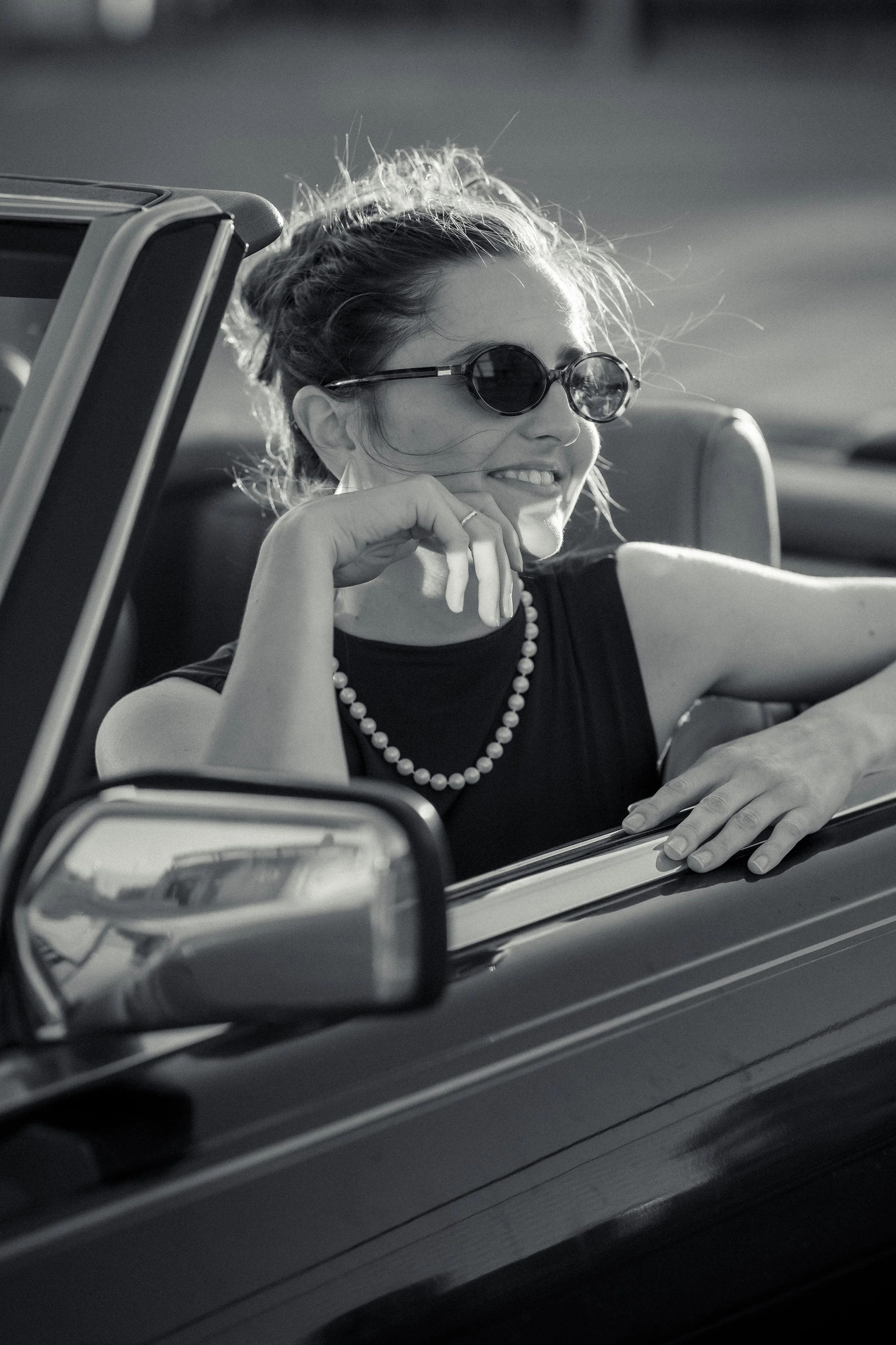 Black and white photo of a woman with sunglasses smiling, seated in a convertible car, with her chin resting on her hand, wearing a pearl necklace and a dark top, with interior car details visible.