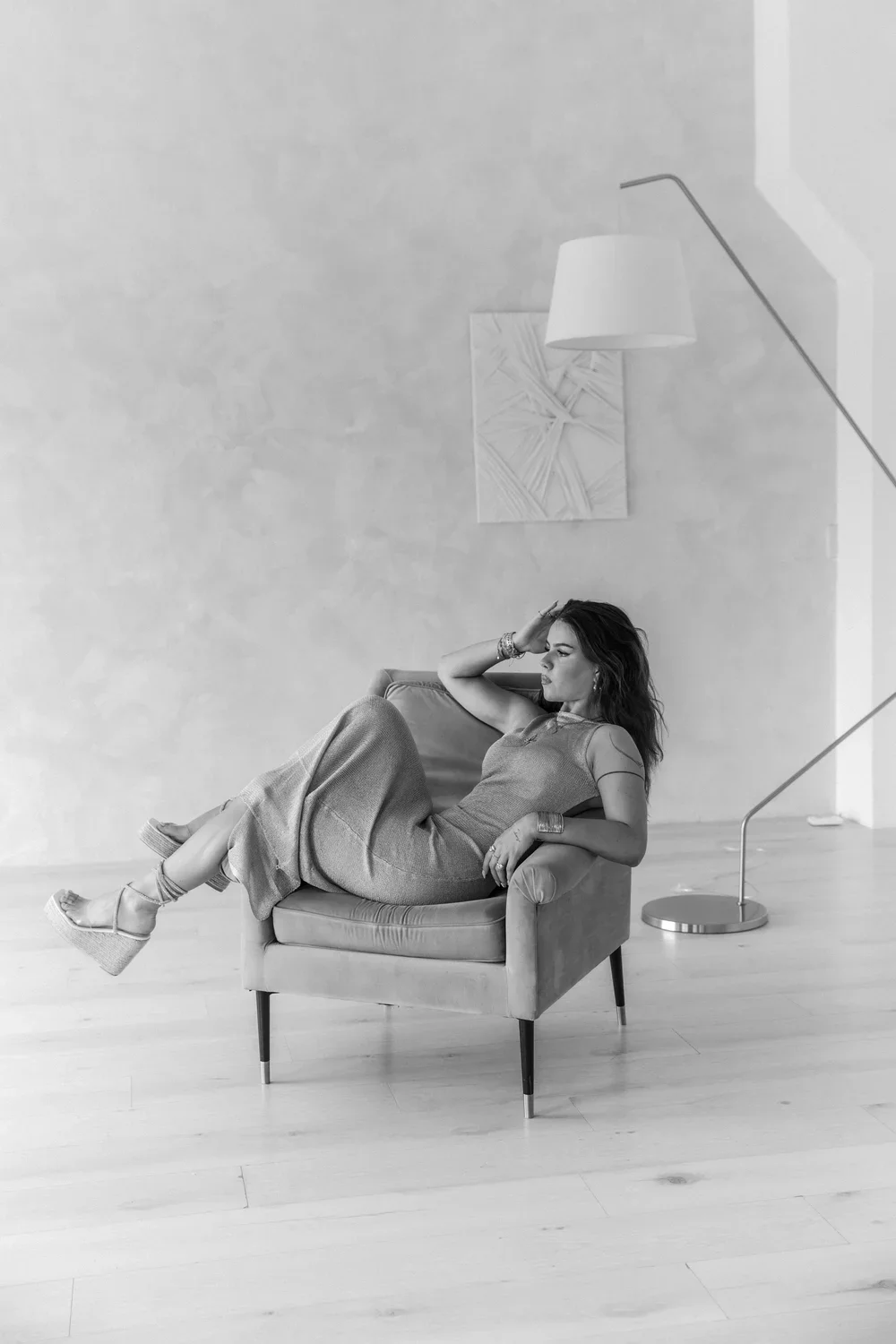 A woman sitting on an armchair in a minimalist room, with one hand resting on her head and the other on her lap, wearing a dress and platform wedge shoes, with a floor lamp and artwork on the wall behind her.