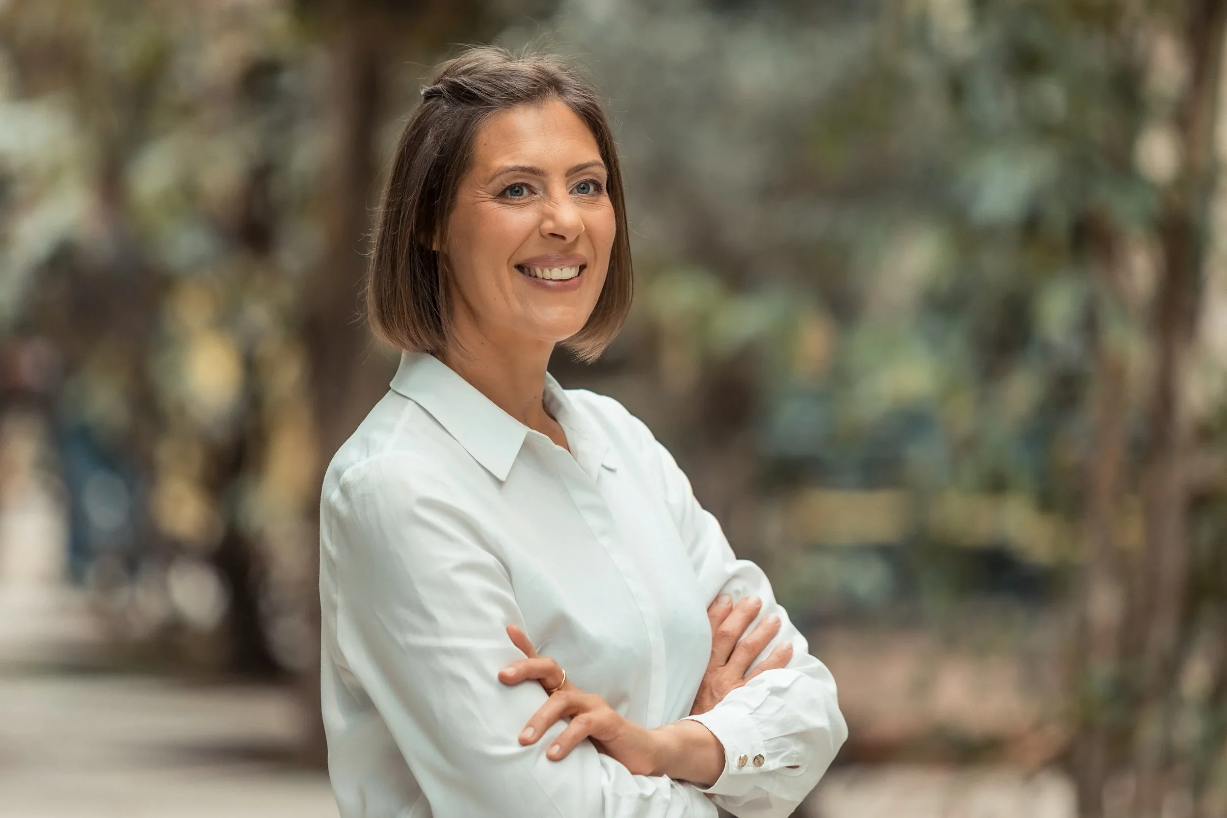 A woman with shoulder-length brown hair and blue eyes standing outdoors with arms crossed, smiling, wearing a white button-up shirt. Blurred trees and foliage in the background.