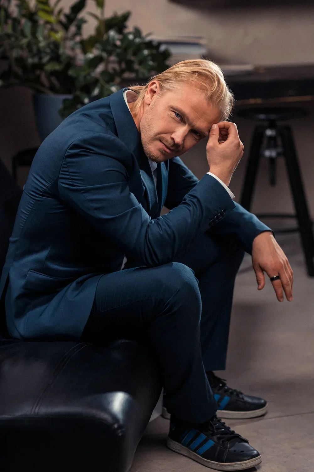 A man with blond hair, wearing a blue suit and black sneakers with blue stripes, sitting on a black leather bench in an indoor setting. He is leaning forward, resting his elbow on his knee, and touching his forehead with his hand, with a serious or c